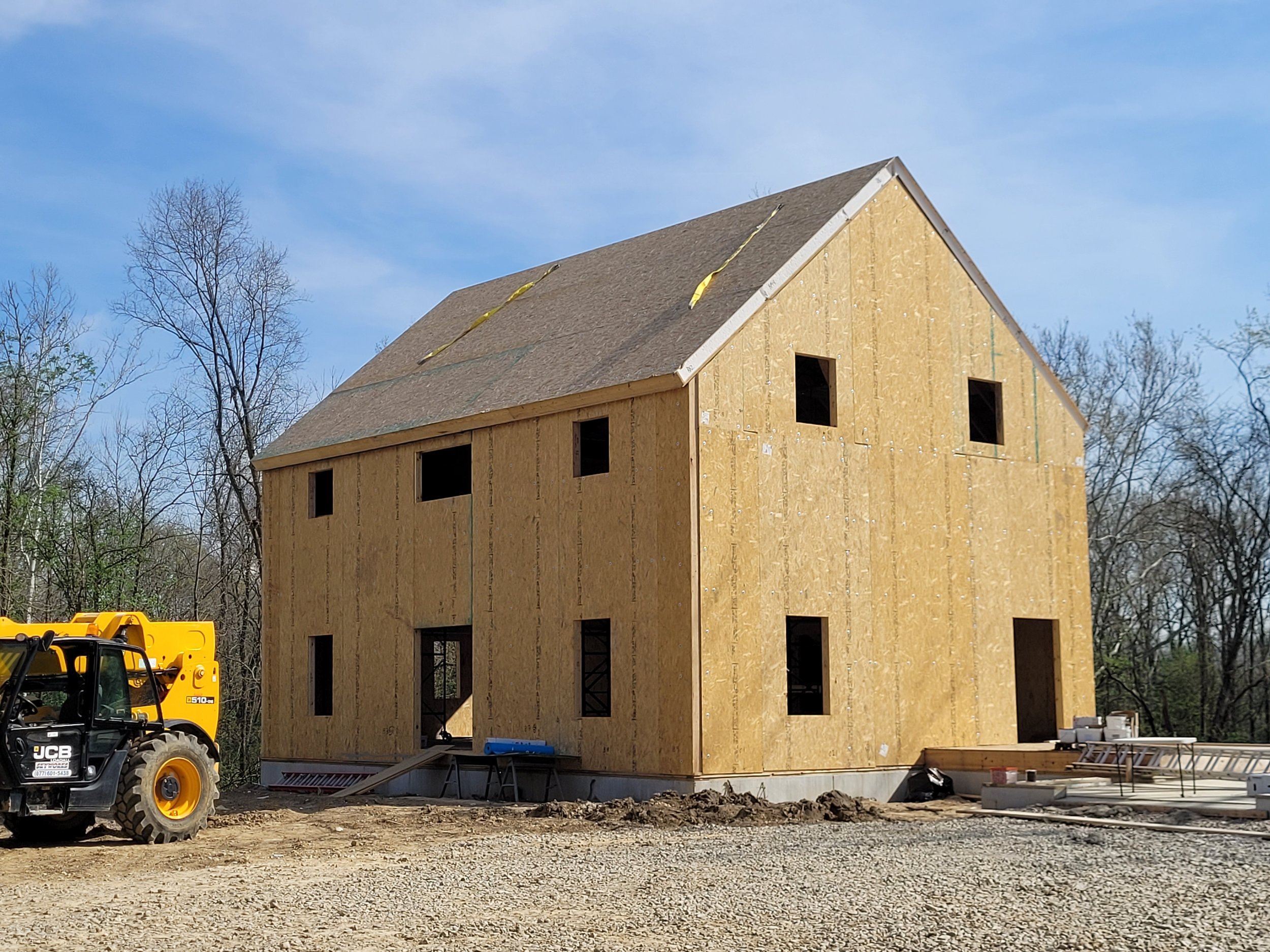 A house under construction with unfinished wooden walls and a roof, surrounded by construction equipment and trees, with a blue sky in the background.