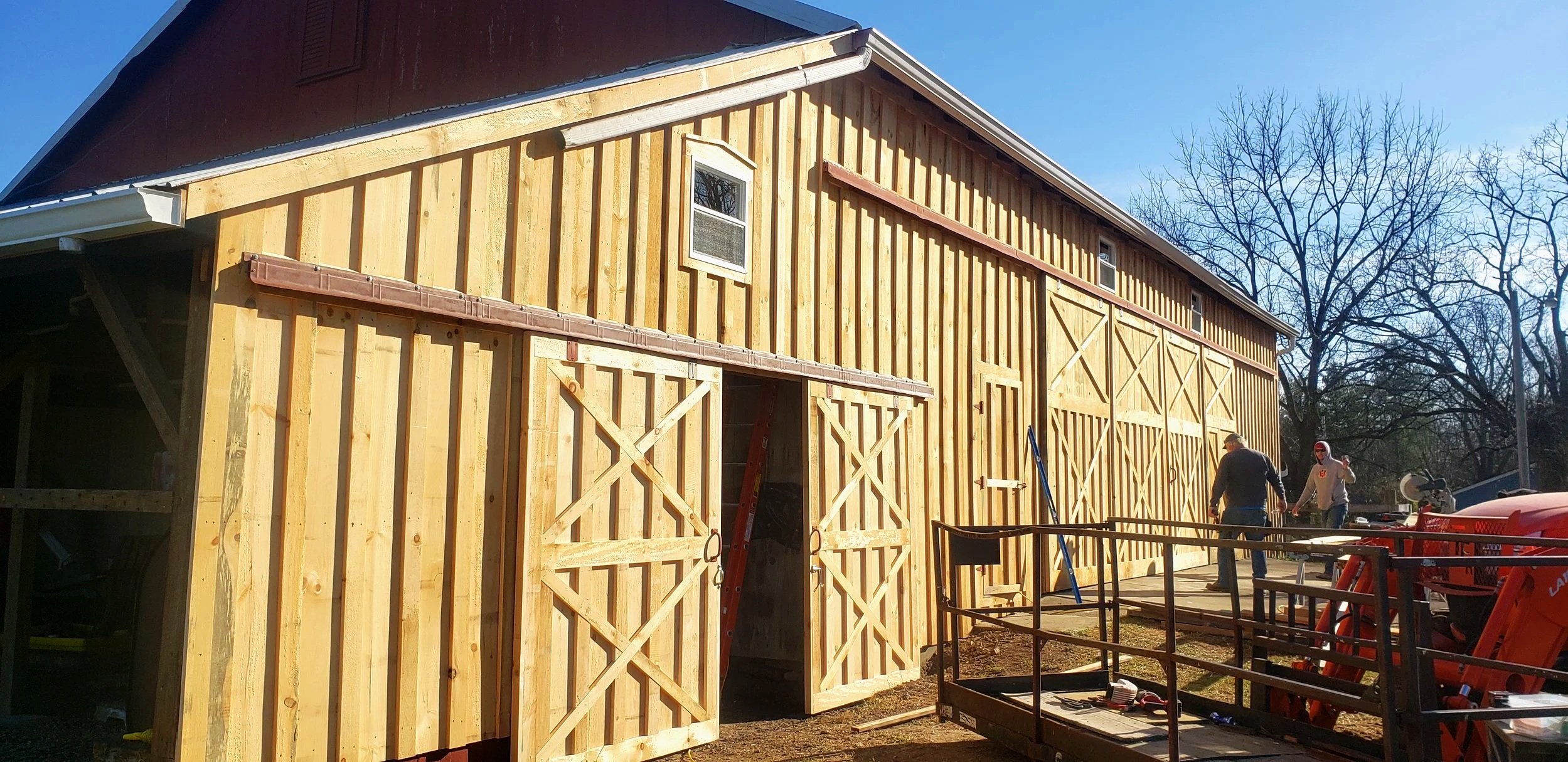 A building under construction with wooden framing, partially installed doors, and two workers on site. The structure is a barn or similar structure, with a partly finished exterior and a clear sky in the background.