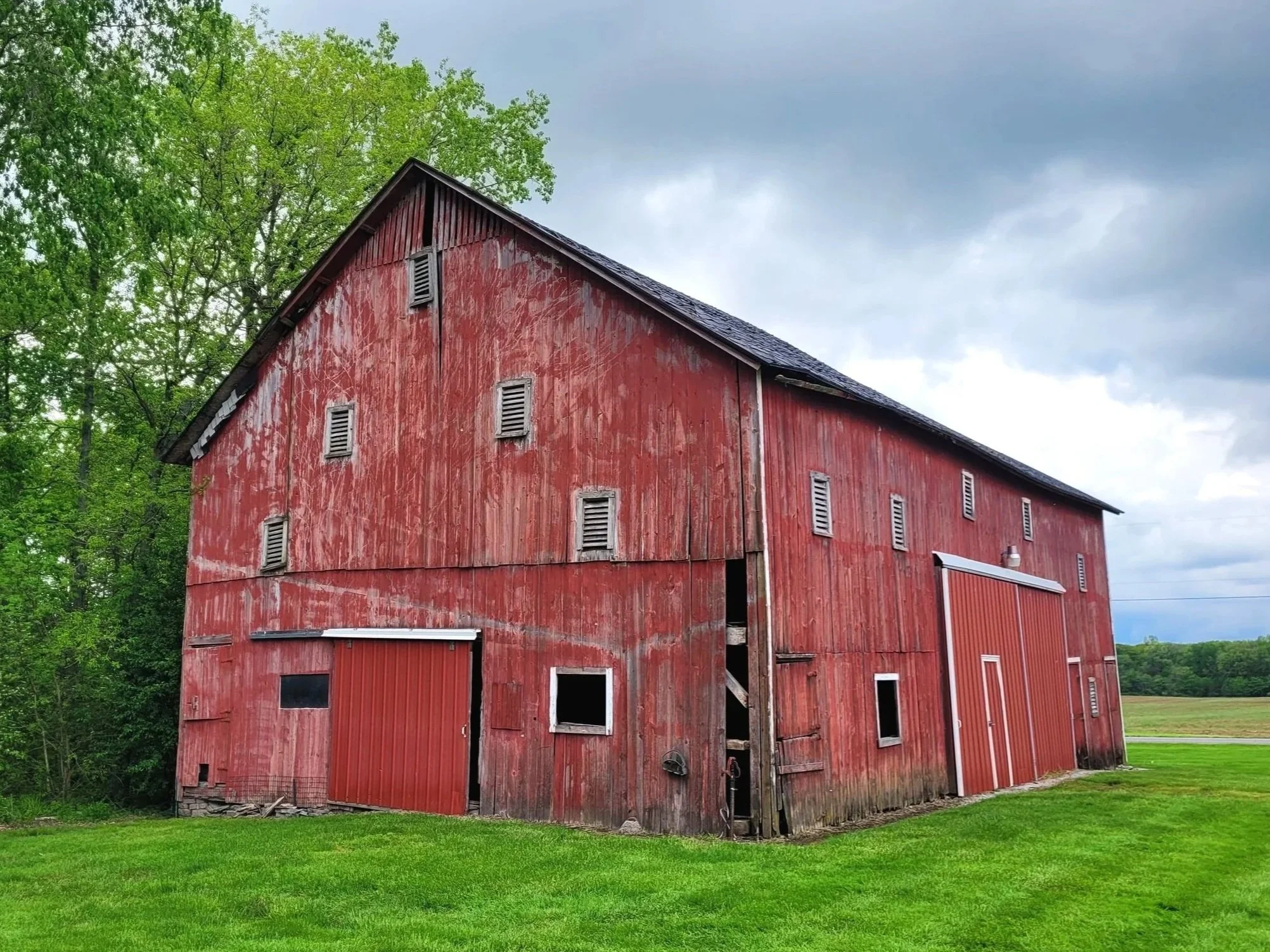Old red barn with weathered wood siding, multiple small windows, and rolling metal doors, surrounded by green grass and trees on a cloudy day.
