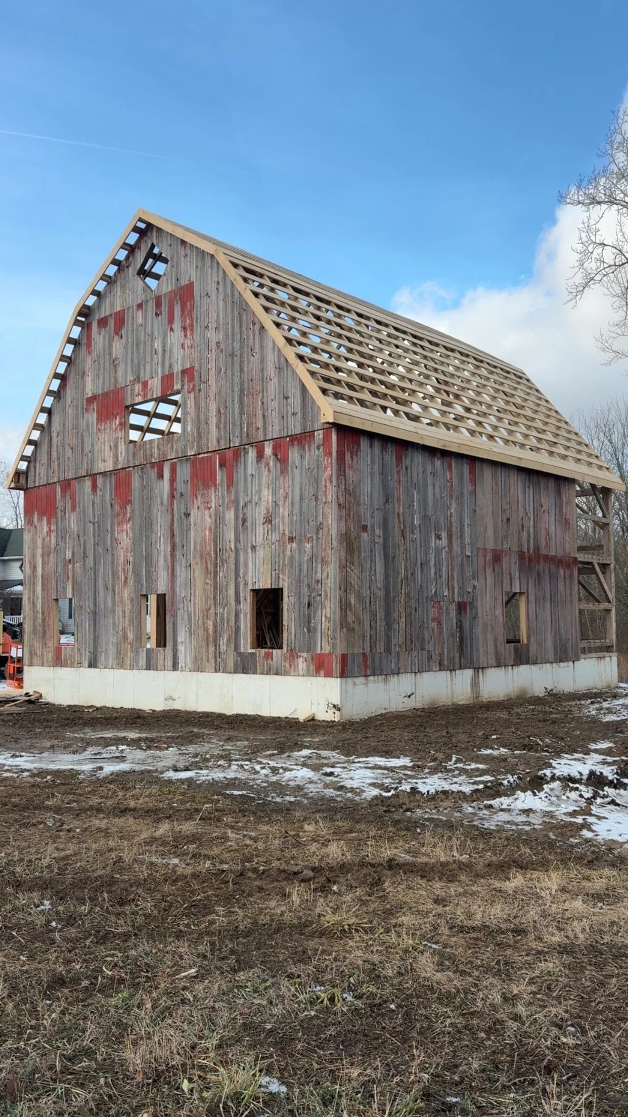A house under construction with wooden exterior walls and a partially built roof, standing on a muddy ground with patches of snow.