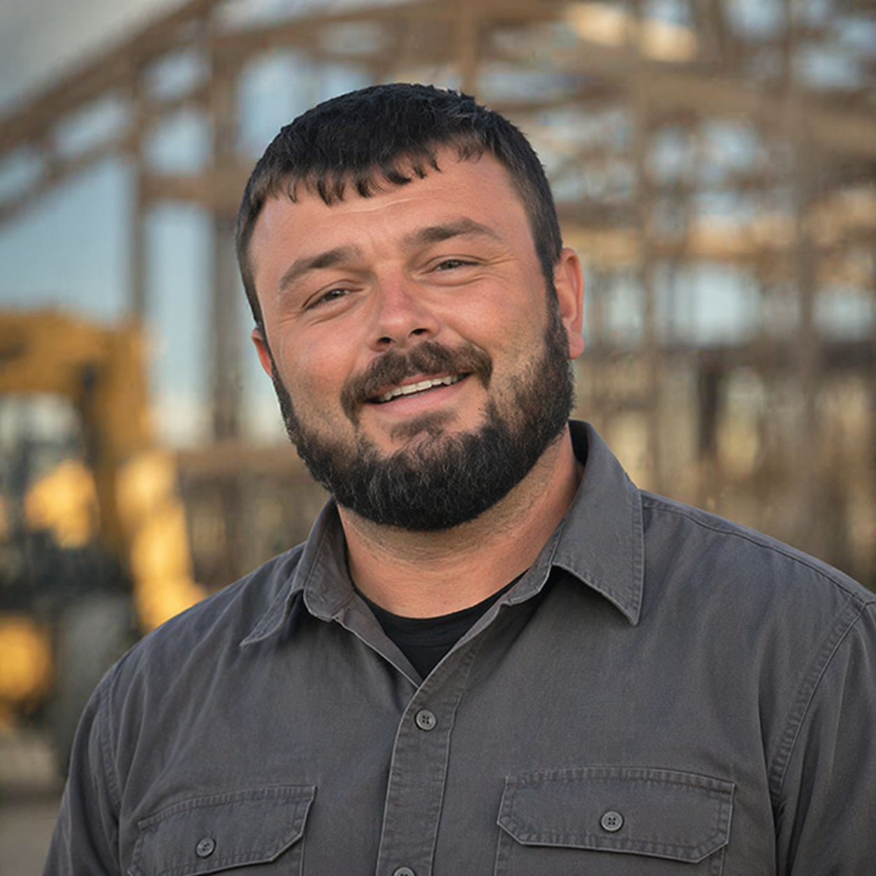 A man with short dark hair and a beard, smiling outdoors with a blurred industrial structure in the background.