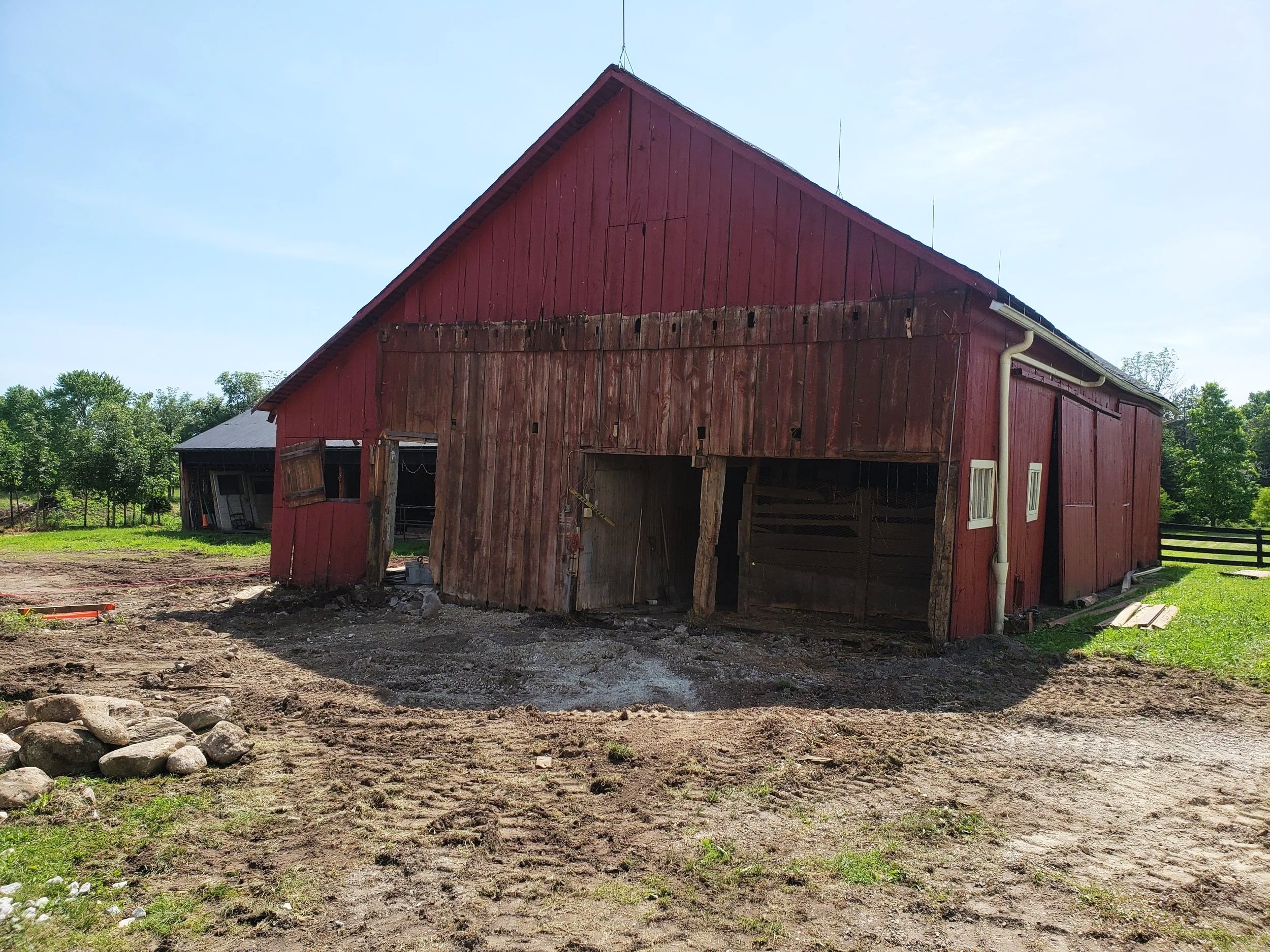 A red barn with weathered wooden siding, some of which is missing or damaged, situated in a rural area with trees in the background. The barn is under construction or repair, with an open doorway and a partly open side, exposing the interior structure. There is dirt ground in the foreground, with some rocks and construction debris.