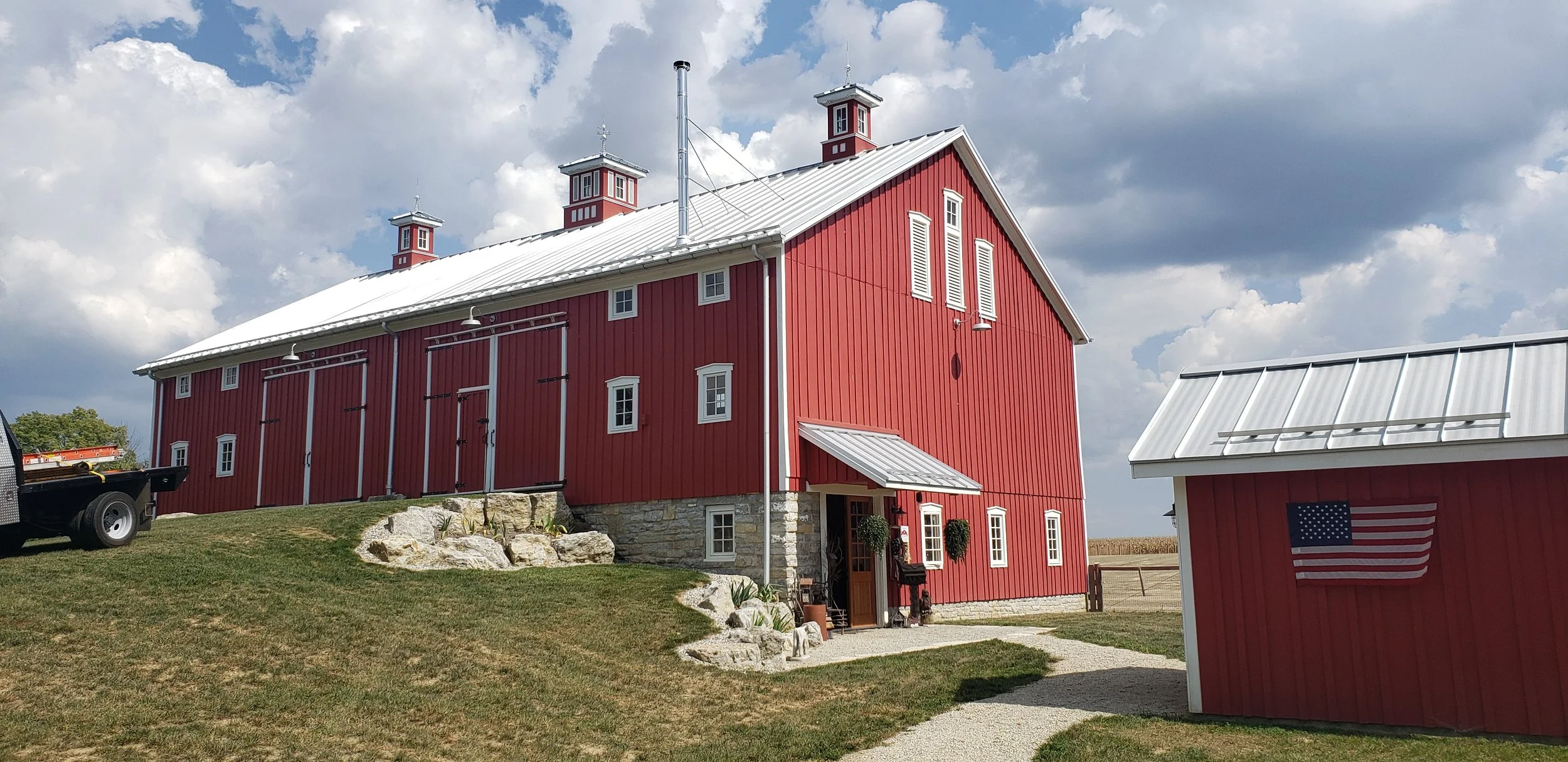 A red barn with white trim and a metal roof positioned in a farm setting under a partly cloudy sky. There is a smaller red structure with an American flag in the foreground on the right side of the image.