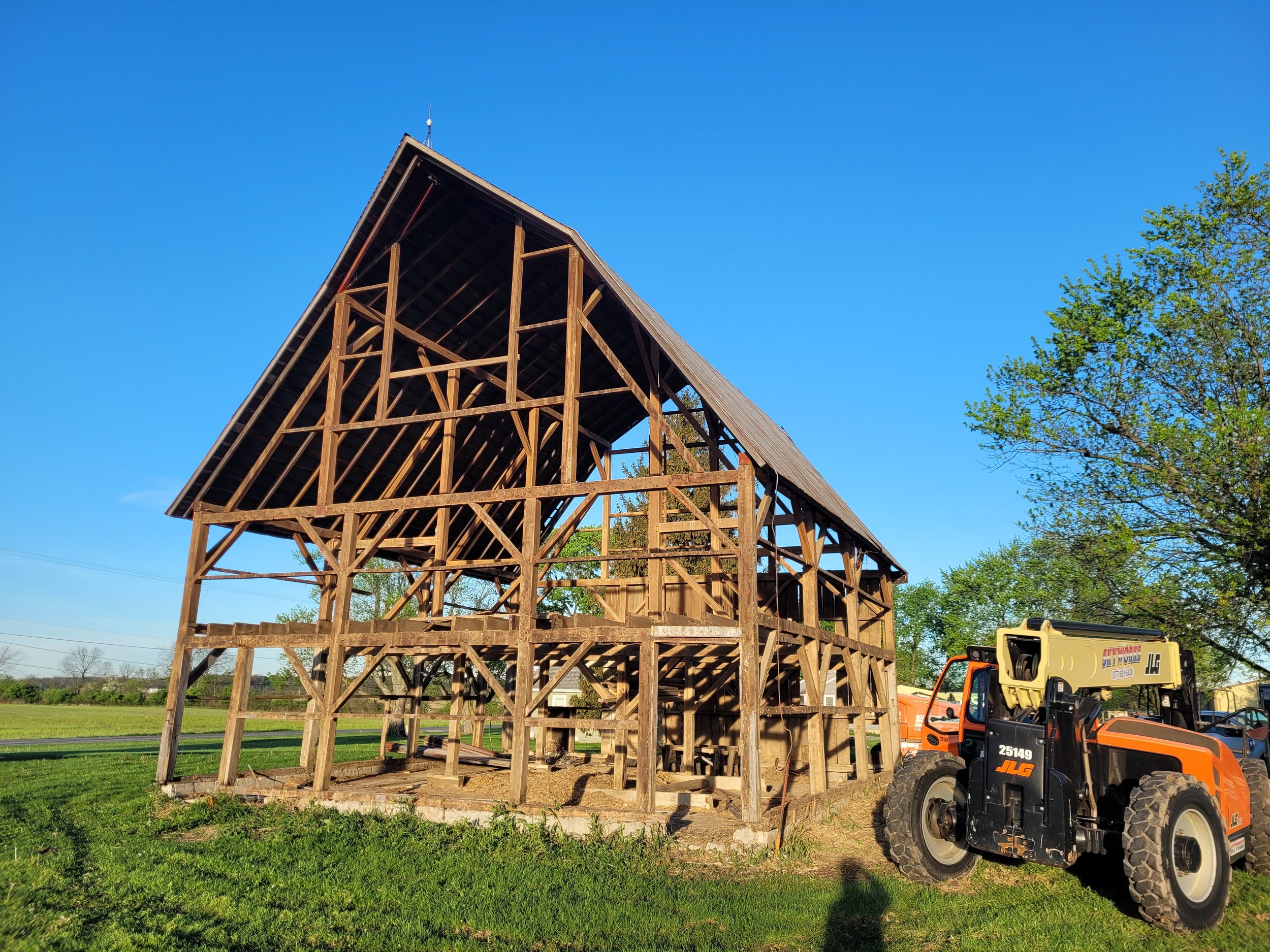 A wooden house frame under construction outdoors on a clear day, with green trees and grass around, and construction equipment nearby.