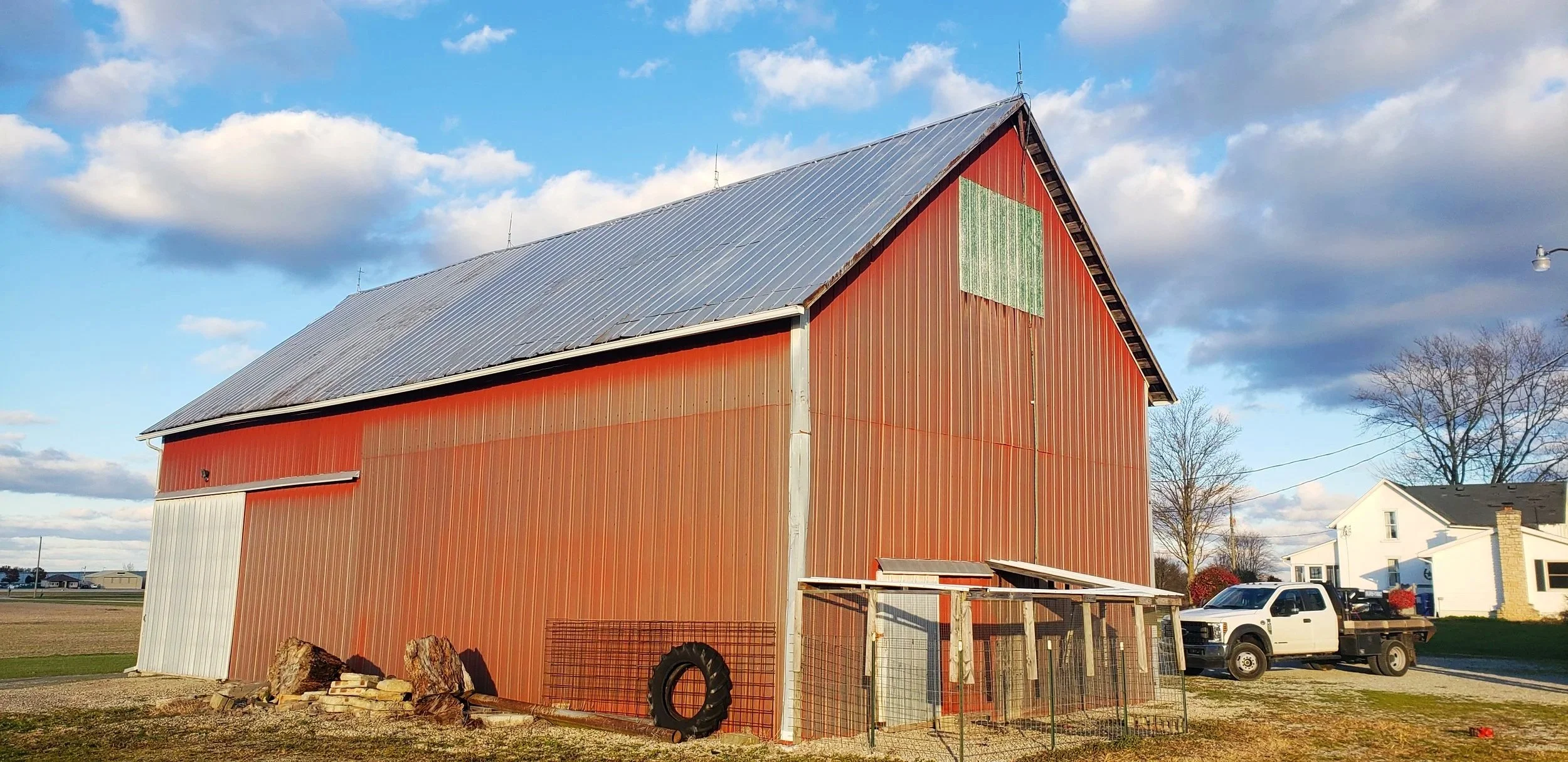 Red barn with a metal roof next to a white house and pickup truck under a partly cloudy sky.