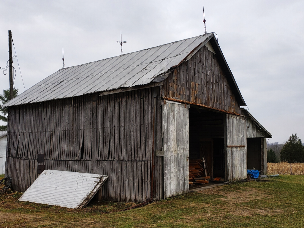 An old wooden barn with a metal roof, weathered wood siding, and open large doors. There are small antennas on the roof and a tilted white panel leaning against the barn. The barn is situated on grassy land with a cloudy sky overhead.