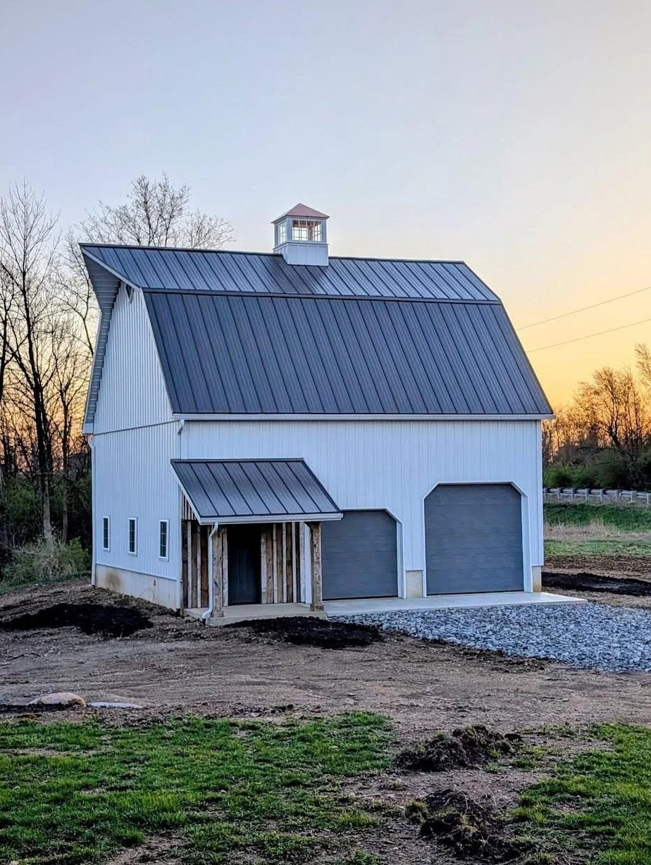 A newly built barn with white siding and a gray metal roof, featuring a small cupola at the top. The barn has two garage doors and a small covered entrance with wooden support beams. The surrounding ground is partly landscaped with grass and gravel.
