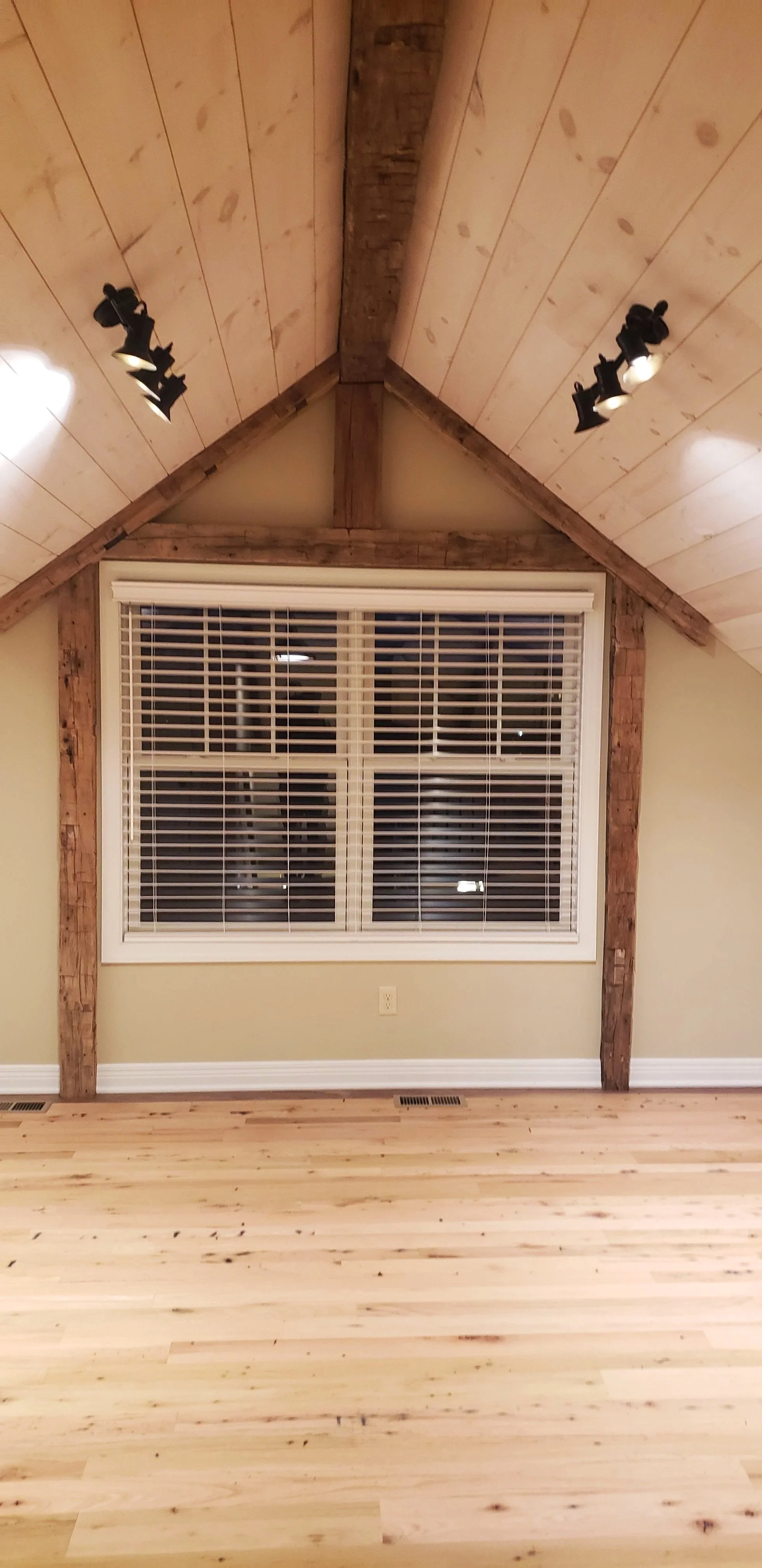 A room with a large window with white blinds, surrounded by exposed wood beams, and a sloped wooden ceiling with track lighting.