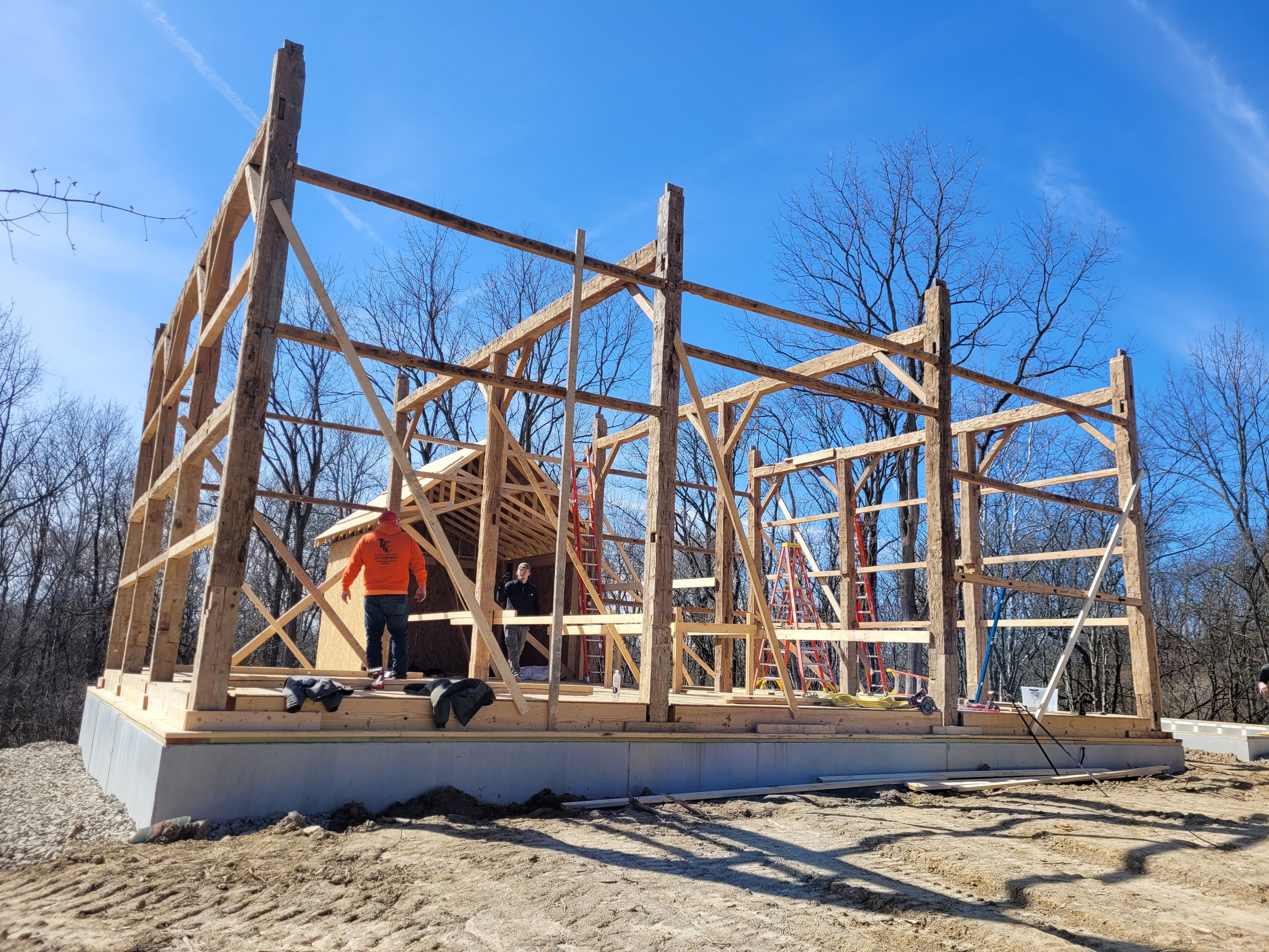 Construction workers building the wooden frame of a house outdoors on a sunny day with leafless trees in the background.