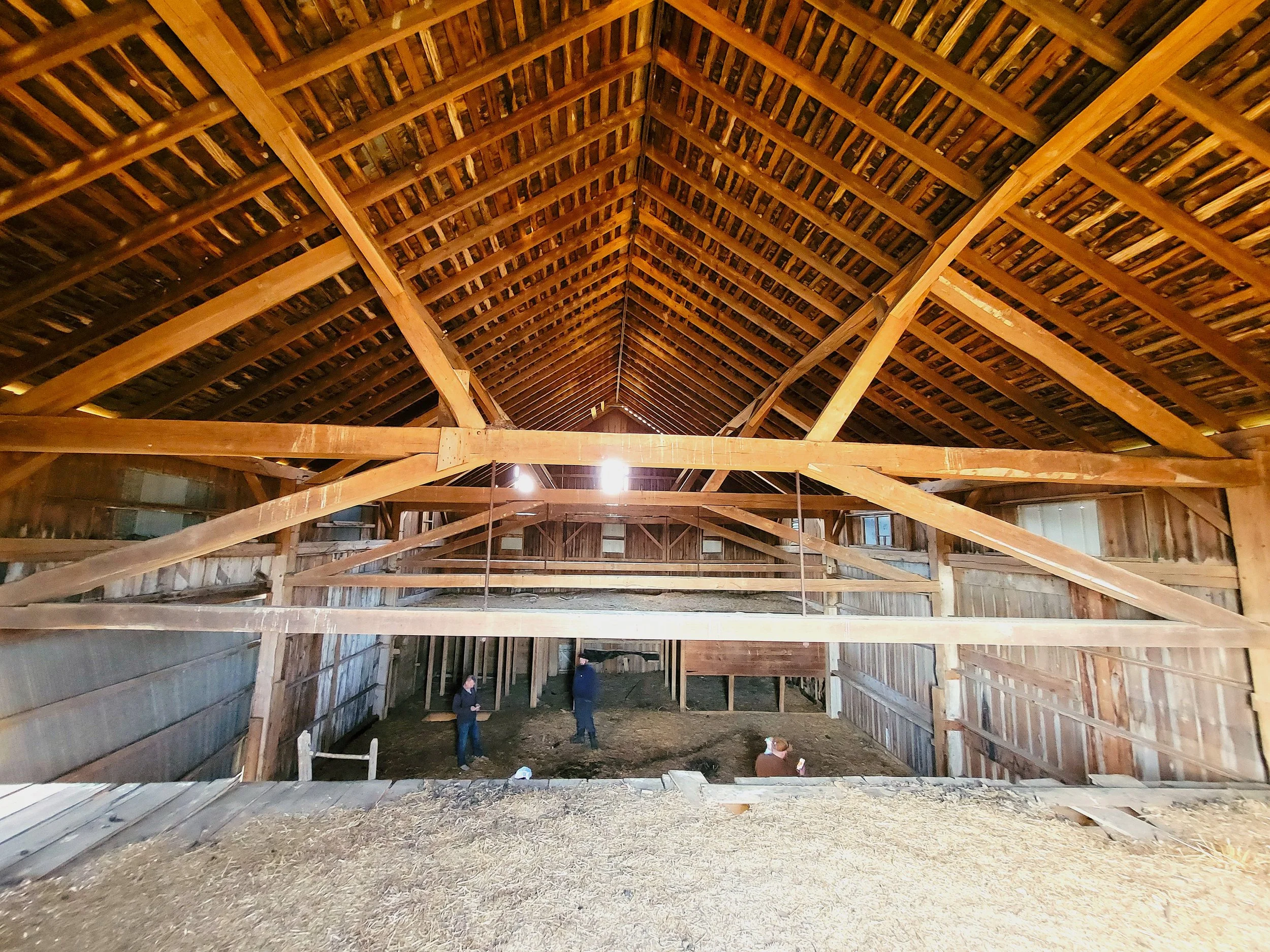 The image shows the interior of a large wooden barn with an open roof structure. There are a few people inside, standing and walking around on a dirt floor. The barn has high ceilings with exposed wooden beams and rafters.