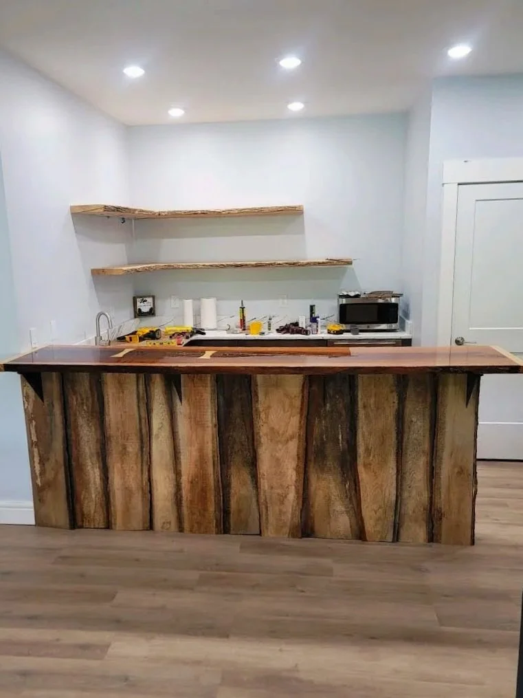Kitchen with a rustic wooden bar counter, light blue walls, two floating wooden shelves, and various small appliances and tools on the back countertop.