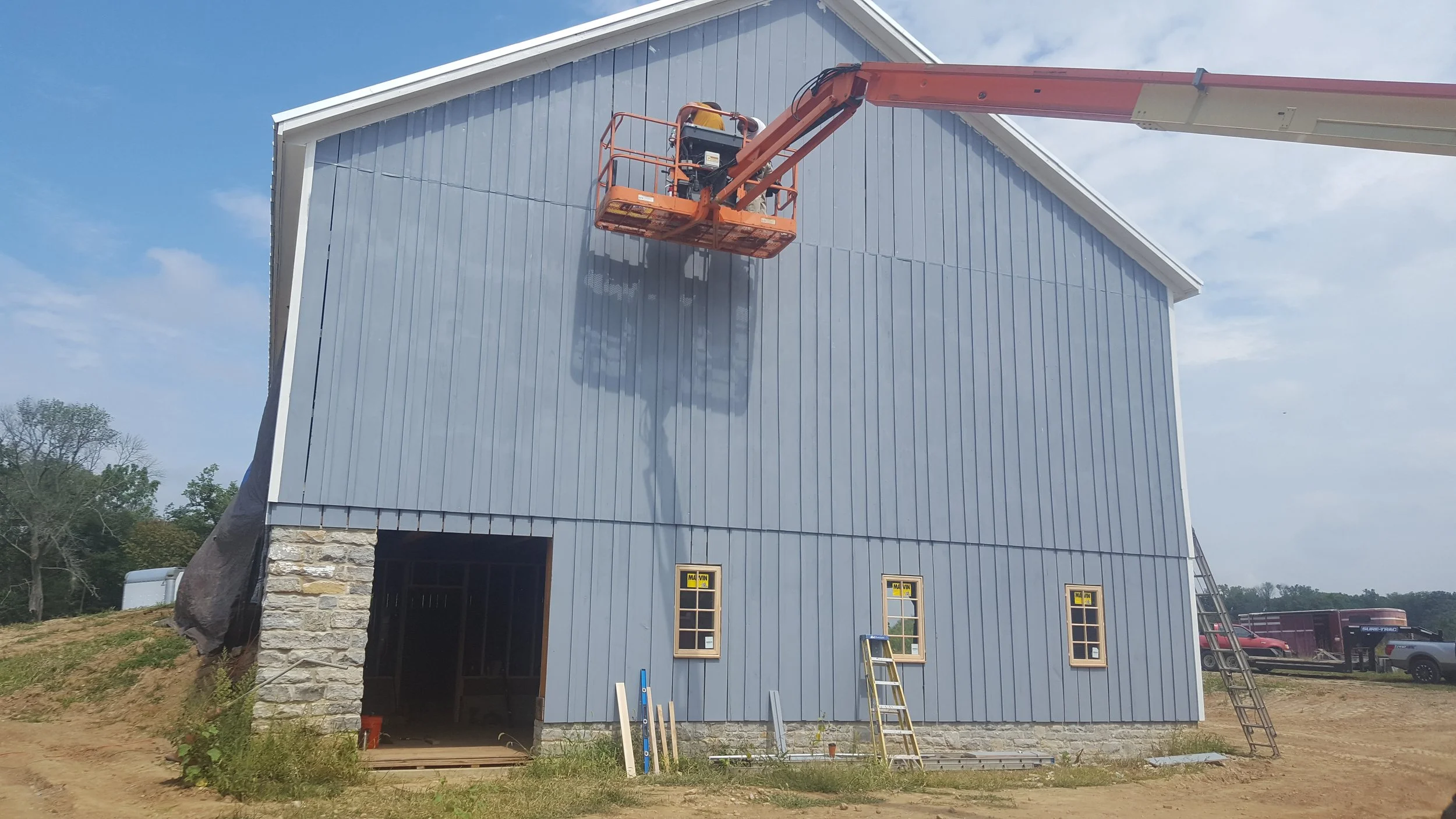 Construction worker using an orange boom lift to work on the upper exterior wall of a house under construction, which is clad in blue siding. The house has a stone foundation and three newly installed windows.
