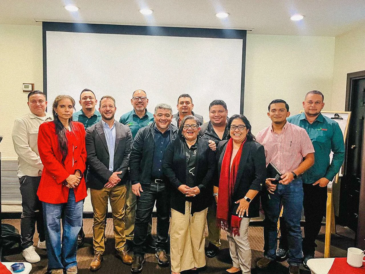 Grupo de personas en una reunión, algunos con vestimenta formal y otros con ropa casual, en un salón con proyector y pizarra.