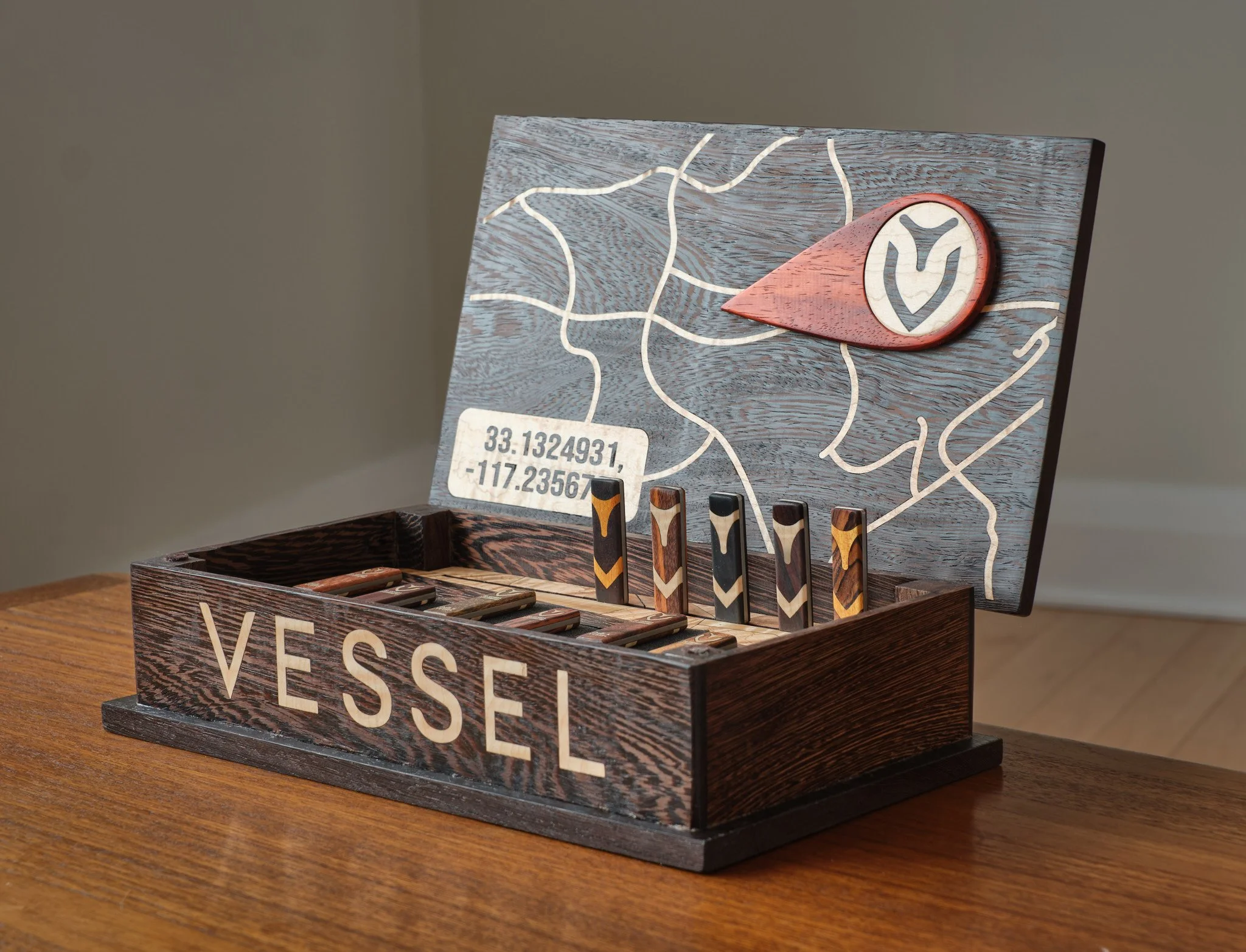 A wooden box with the word 'VESSEL' printed on the front, containing billiard cue sticks. The inside of the lid features a map design with coordinates 33.1324931, -117.23567 and a logo with a leaf.