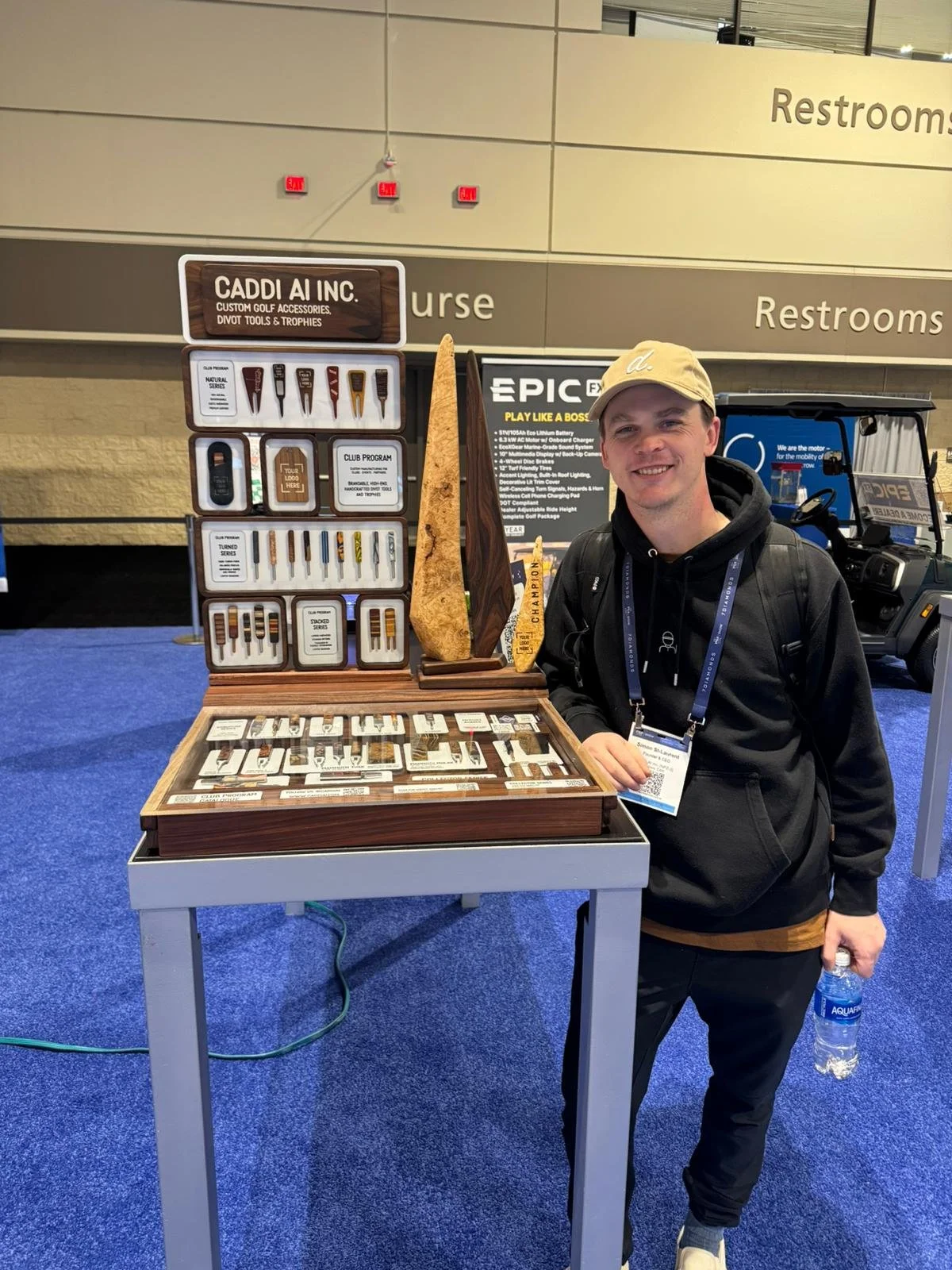 A smiling man in a beige cap, black hoodie, and black pants standing next to a display of golf accessories at a convention or trade show. The display includes various golf tools and trophies, with a large wooden surfboard as a centerpiece.