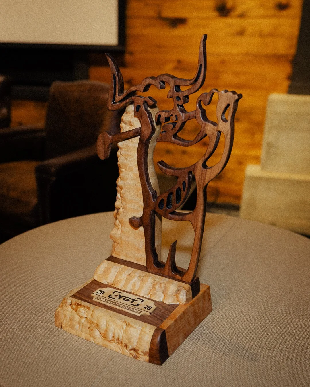 A wooden trophy featuring a cut-out model of a deer with antlers. The trophy has a wooden base with engraved text and a smaller metal plaque.