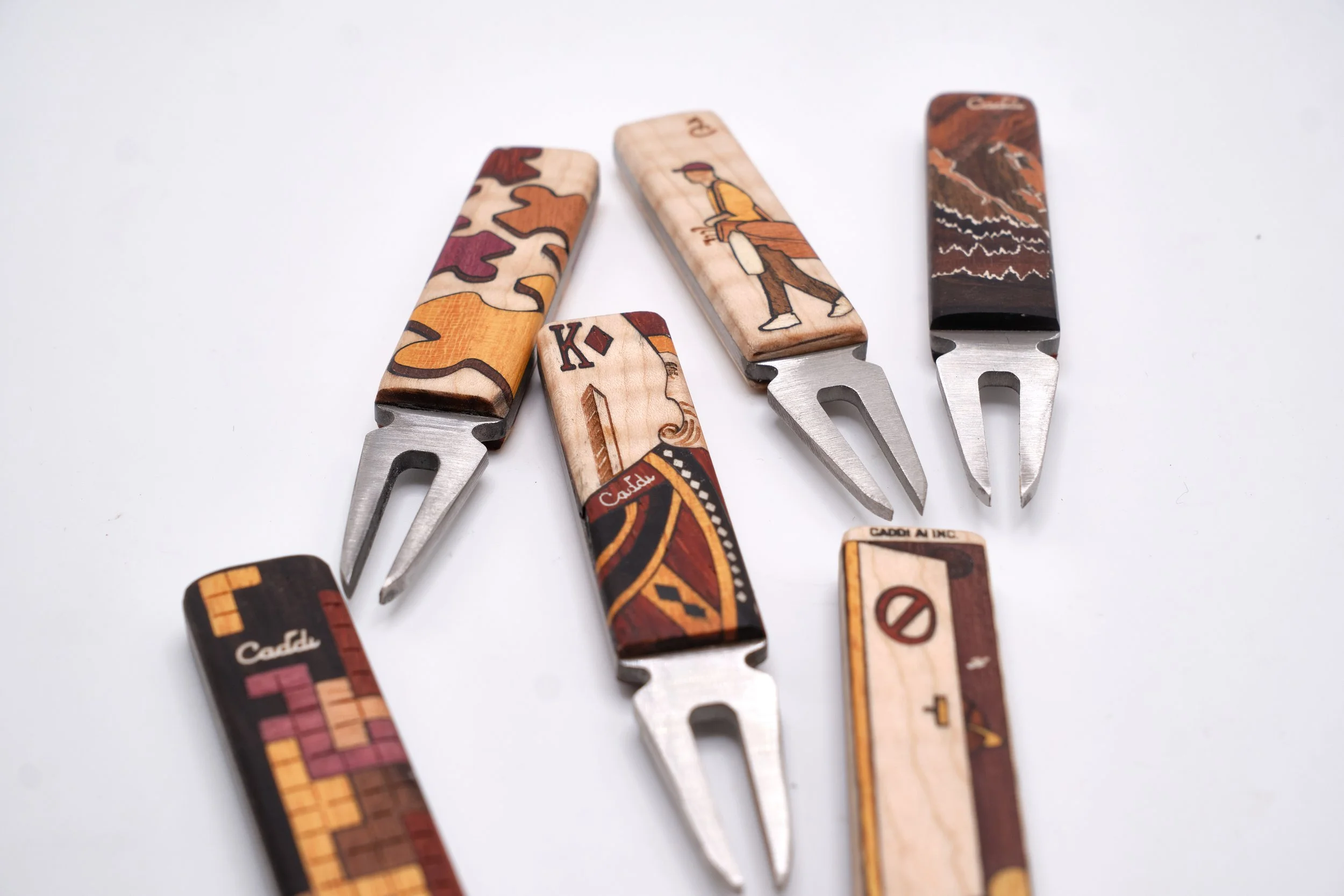 Collection of wood-ended forks with decorative designs, including a mountain scene, artwork, and text, arranged on a white surface.