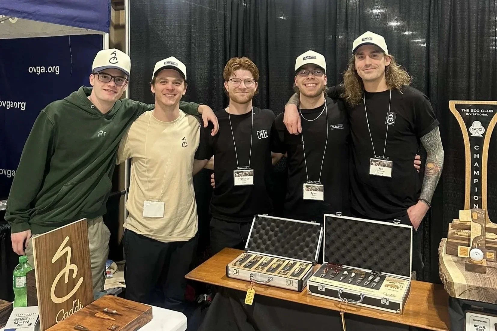 Five young men standing together at a trade show booth with golf merchandise, some wearing hats and name tags, with cases of golf clubs and wooden signs on display.
