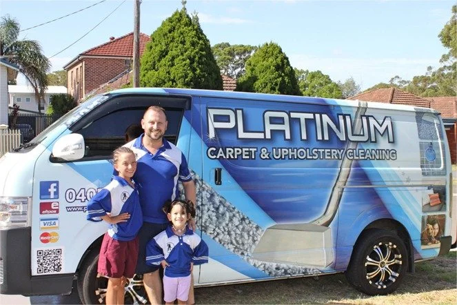 A family of three standing next to a large blue van with "PLATINUM CARPET & UPHOLSTERY CLEANING" branding parked on a residential street.