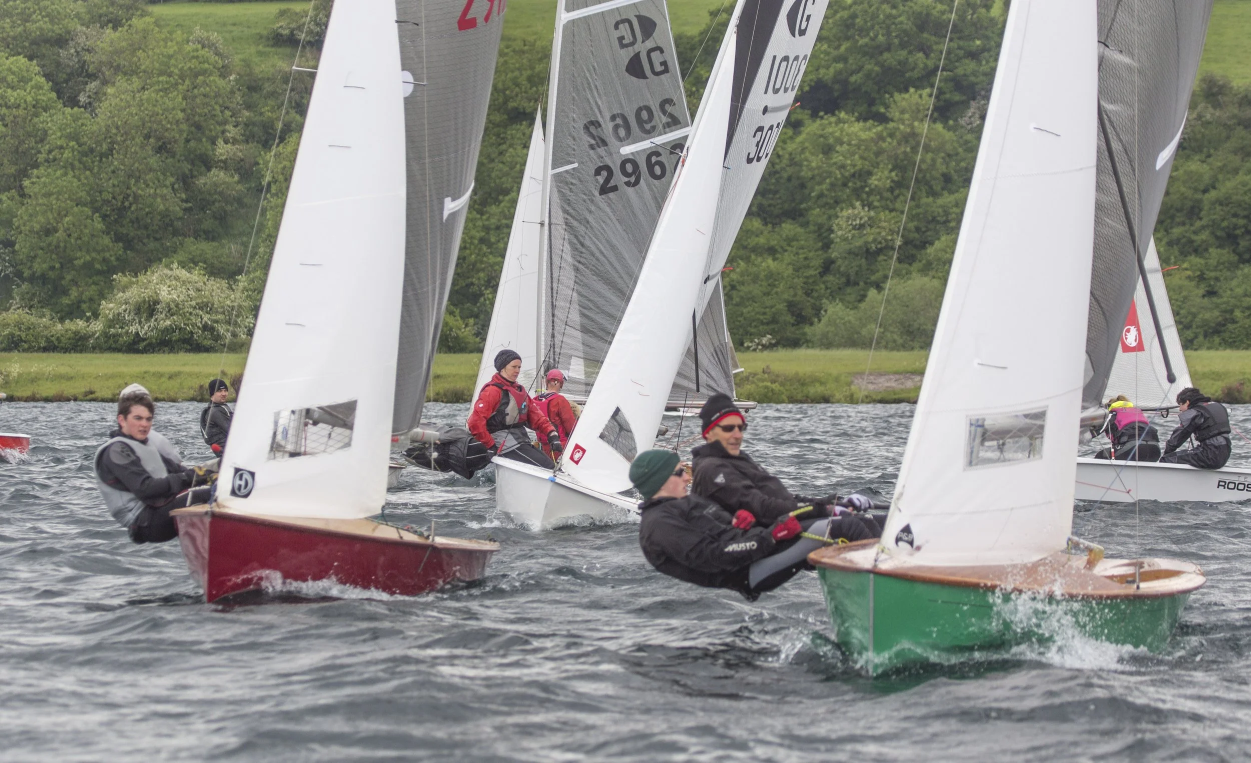 Sailing race with multiple sailboats on choppy water, sailor crews in jackets and hats, green trees and grassy shoreline in background.