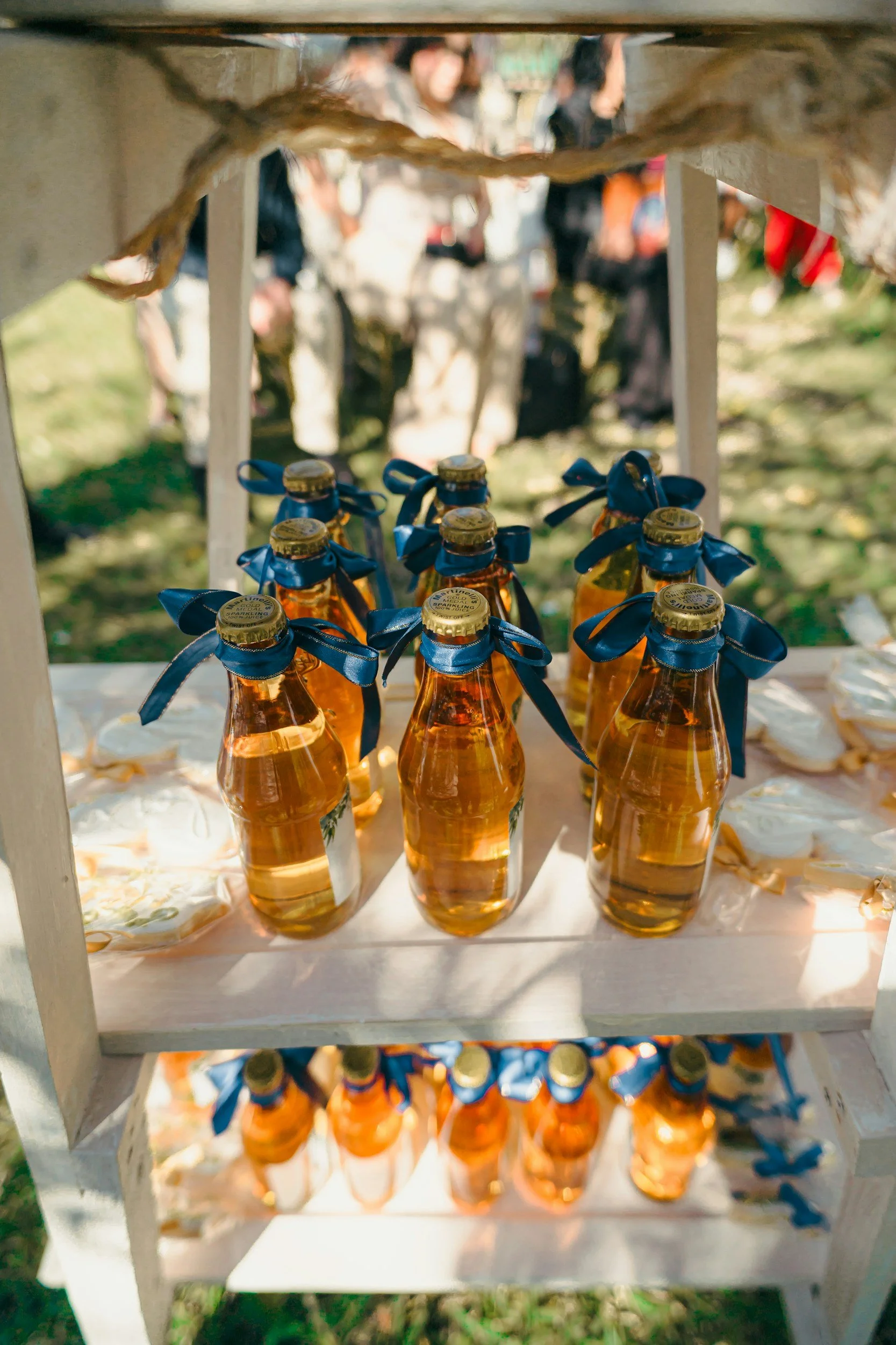 A white wooden shelf with several small bottles of honey, each with a blue ribbon tied around the neck, placed on a white cloth. People are blurred in the background, possibly at an outdoor market or event.