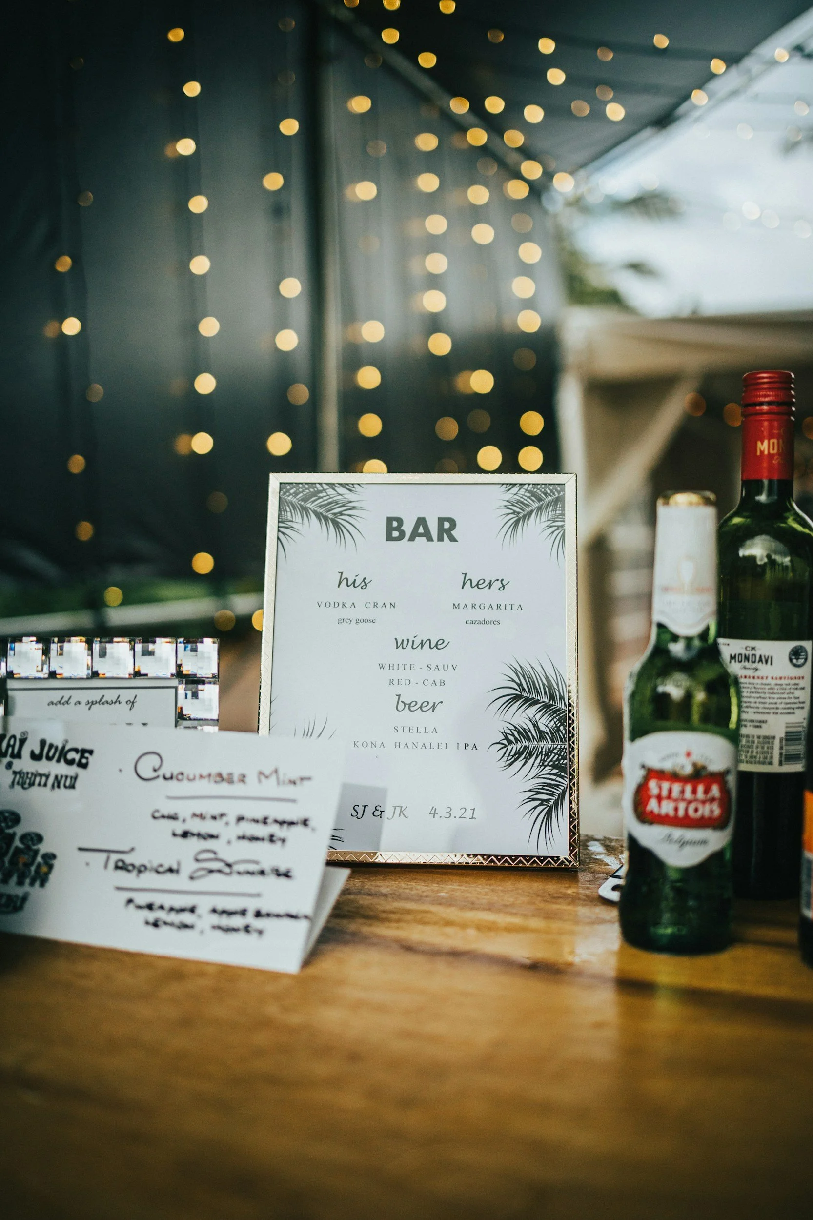 A bar menu on a white paper frame with a tropical design, listing drinks such as vodka cran, margarita, white sauv, red cab, Stella beer, and Kona Hanalei IPA. In the background, there are bottles, a blurred string of yellow fairy lights, and handwritten notes on separate papers on a wooden table.