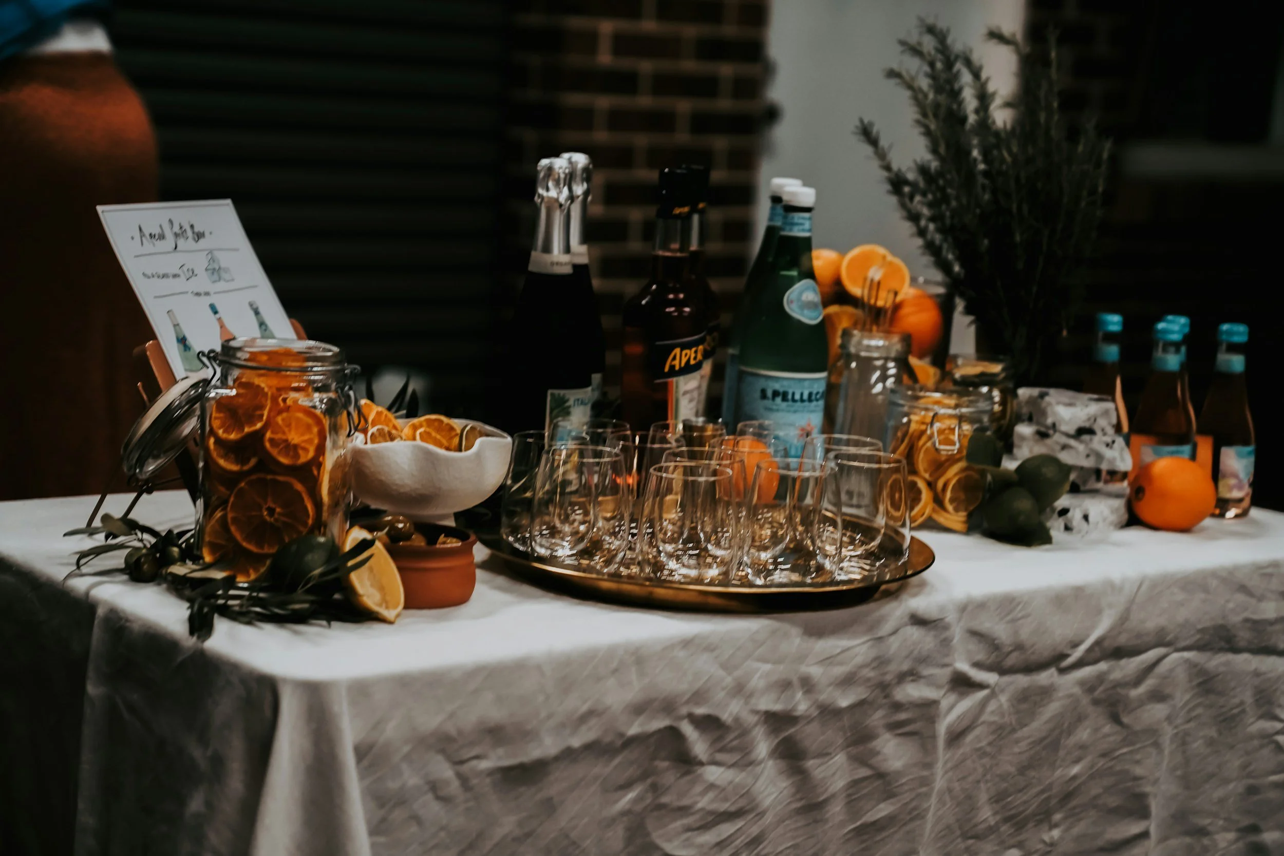 A table with a white tablecloth featuring a display of sliced oranges, green limes, and a jar of dried orange slices. Behind the setup, there are bottles of sparkling water, soda, and other beverages, along with a potted plant and a sign with illustrated bottles and the words 'good life.'
