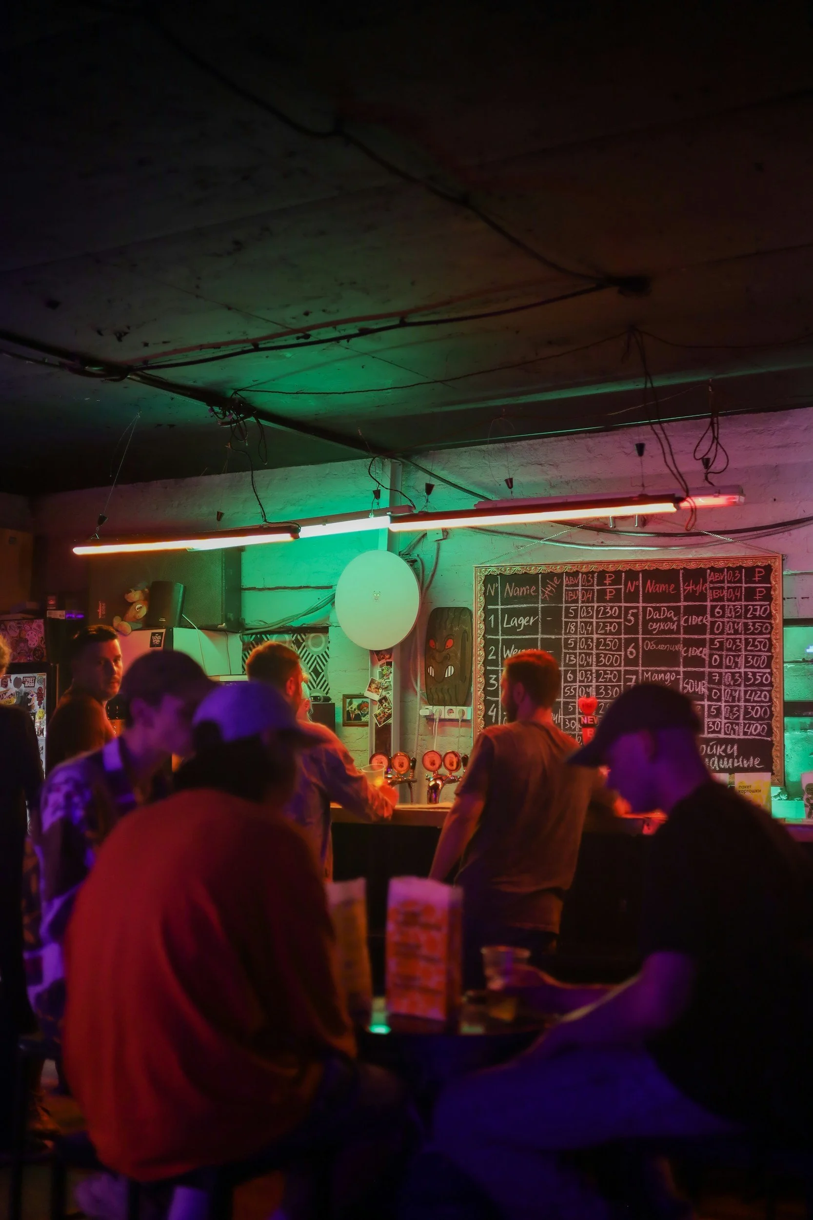 People gathered in a dimly lit bar with neon lights and a chalkboard menu on the wall.