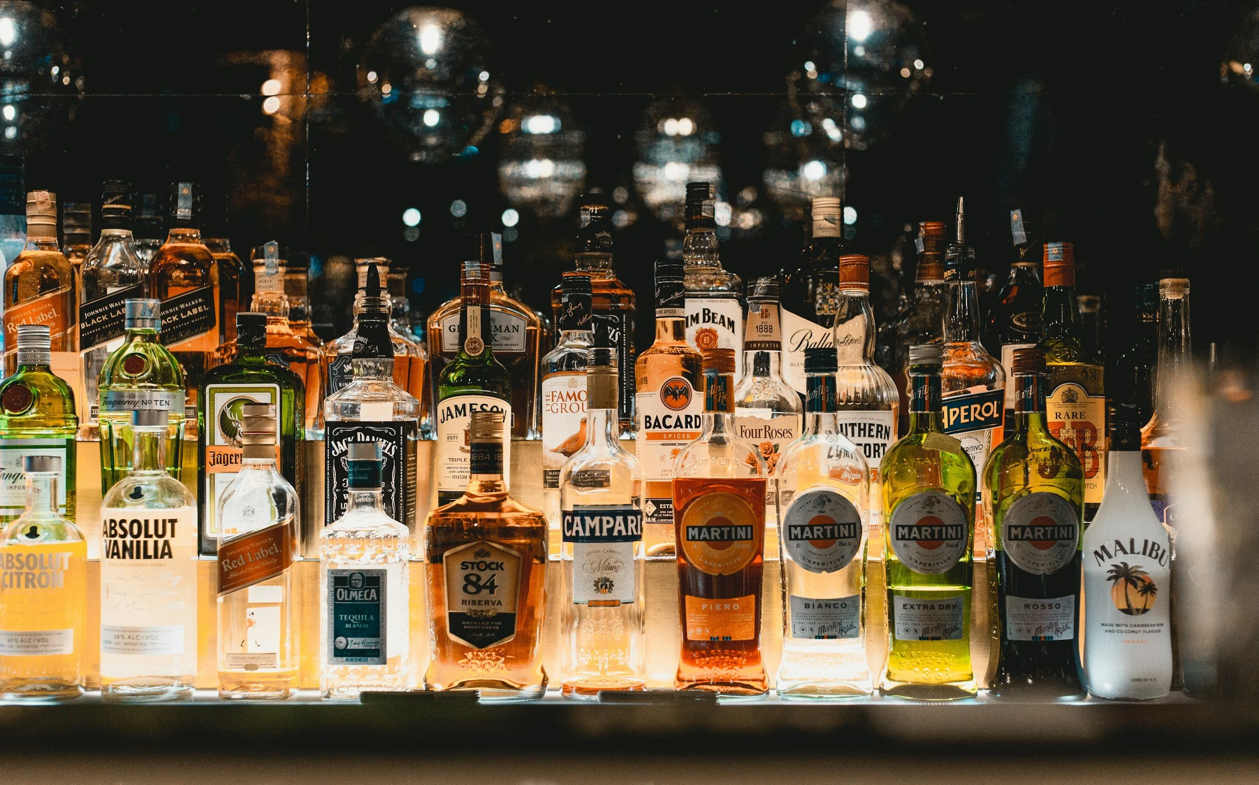 A variety of liquor bottles on a backlit bar shelf, including brands like Absolut, Tanqueray, Jameson, Bacardi, Martini, Malibu, and others, arranged in multiple rows.