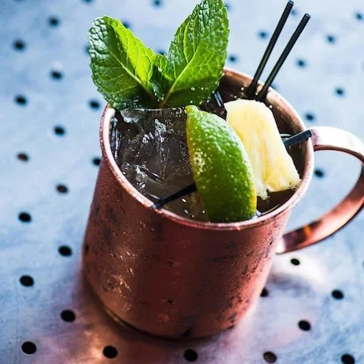 A copper mug filled with a dark beverage, garnished with fresh mint leaves, a lime wedge, and a pineapple chunk, with two black straws.