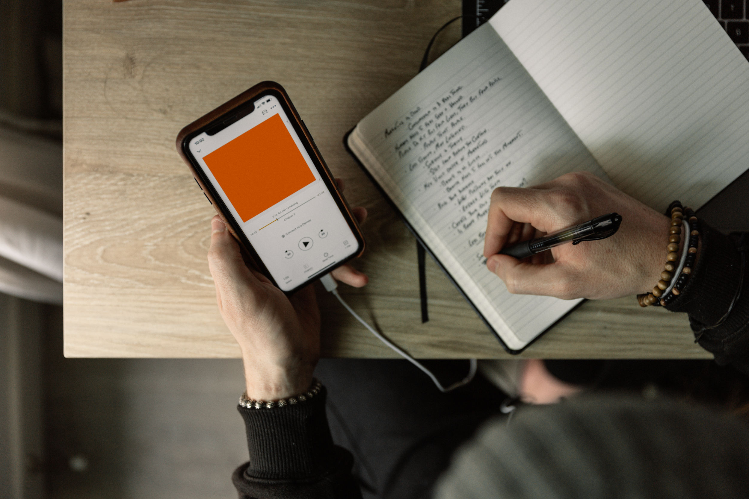 A person holding a smartphone playing audio, while writing notes in a notebook with a black pen on a wooden desk. The person is wearing bracelets on their wrist.
