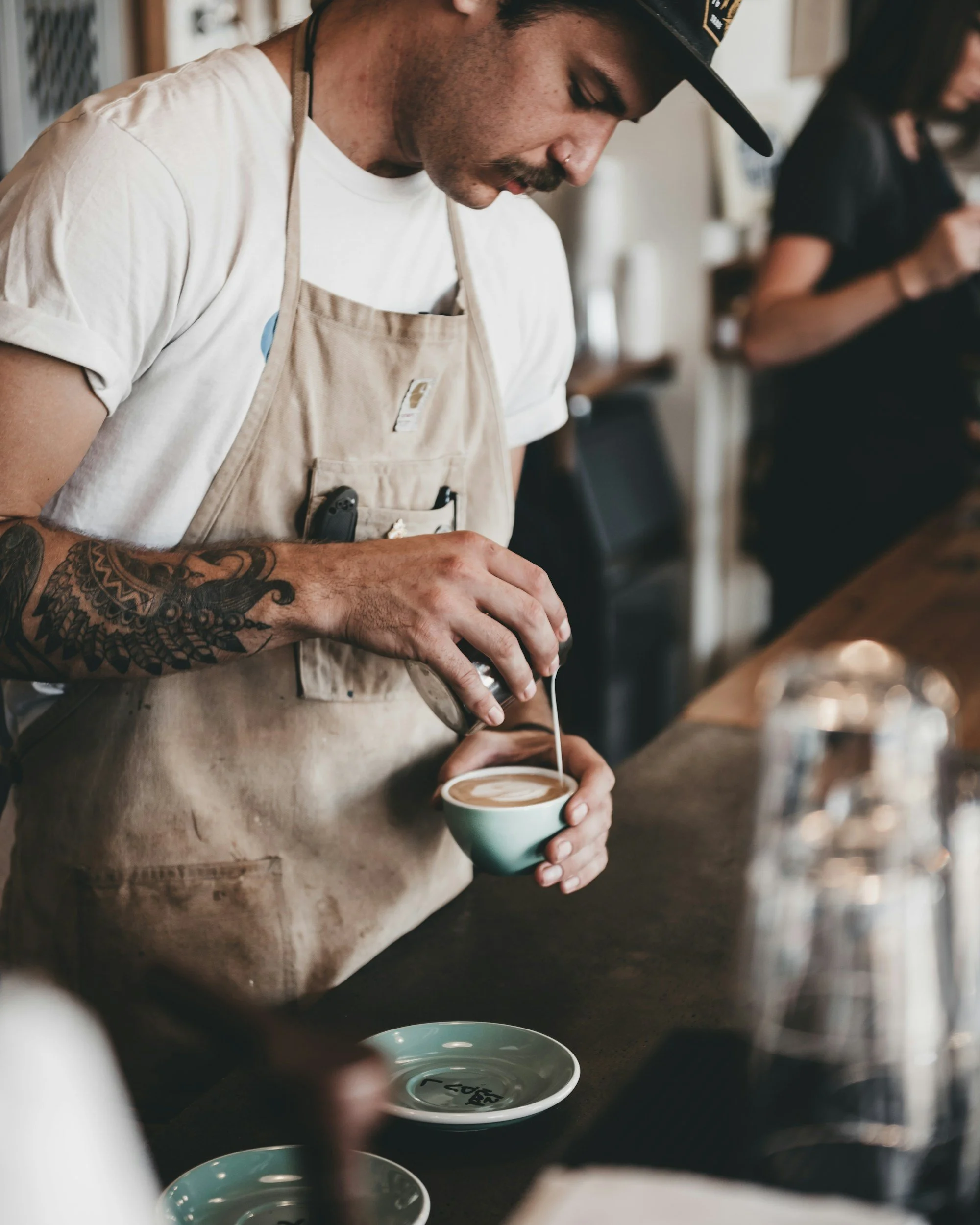 Barista with tattooed arm pouring steamed milk into a coffee cup in a coffee shop.