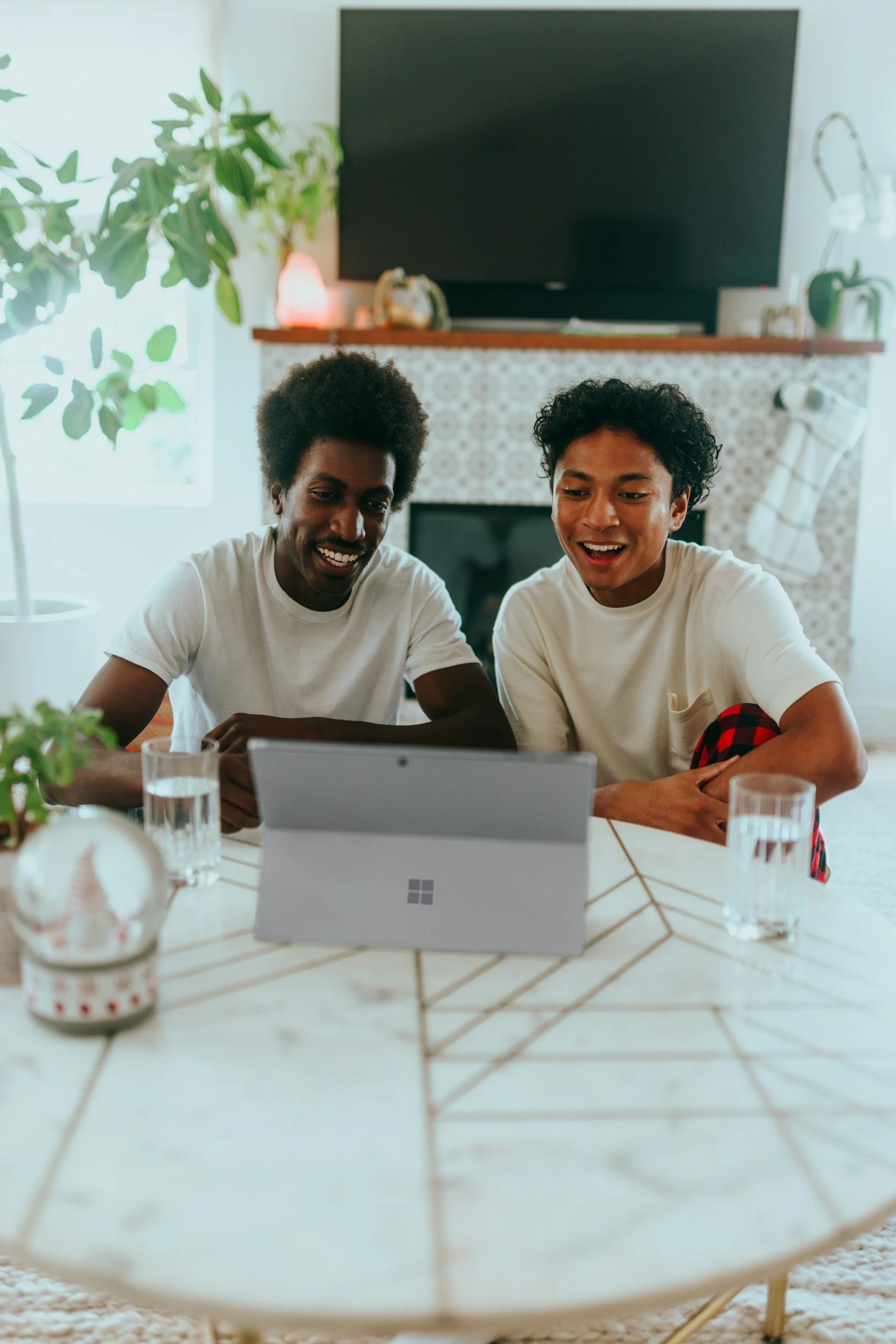 Two young men with dark and curly hair smiling and looking at a tablet while sitting at a round dining table in a living room decorated with plants, a fireplace, and a TV.