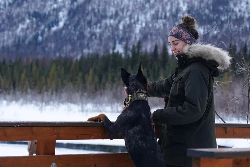 A woman in a winter coat with a fur-lined hood and a headband is smiling at a black and tan dog with a collar, who has its front paws on a wooden railing, looking out over a snowy landscape with trees and a frozen body of water.