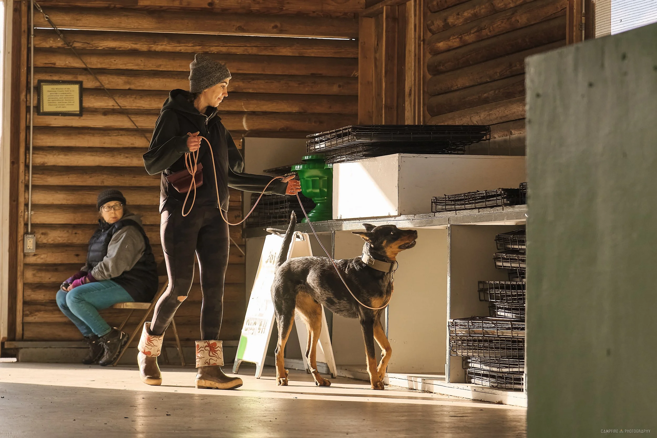 Two women indoors, one standing with a leash attached to a black and tan dog, and the other sitting on a bench. The woman standing is wearing a gray beanie, black jacket, and patterned boots, while the woman sitting is wearing glasses, a black vest, and teal pants. The setting appears to be a wooden interior, possibly a pet-friendly space or cabin.