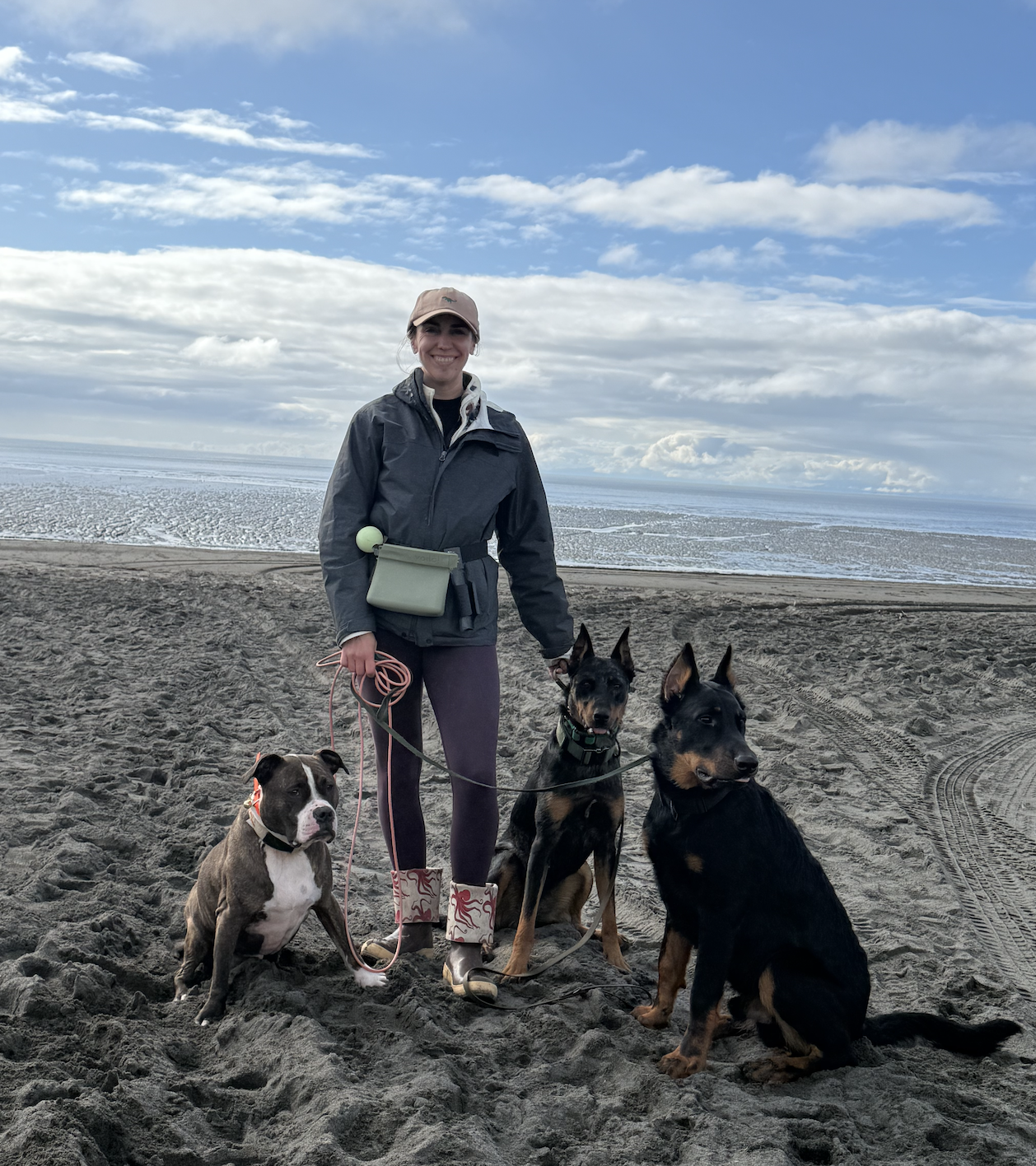 A woman standing on a sandy beach with three dogs, smiling, with a cloudy sky and body of water in the background.