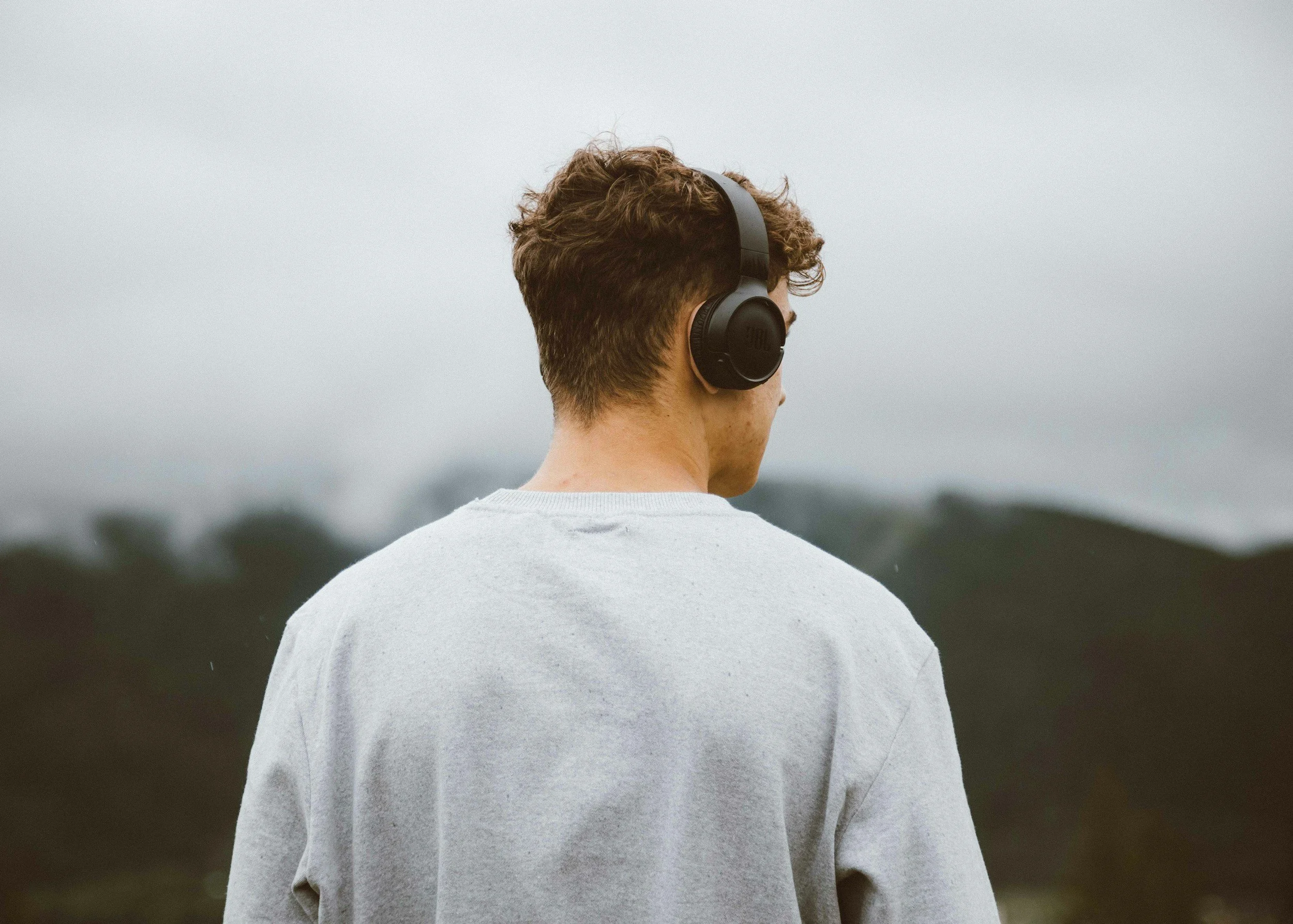 Back view of a young man with short curly hair wearing a white sweatshirt and black headphones, standing outdoors with a cloudy sky and blurred landscape in the background.