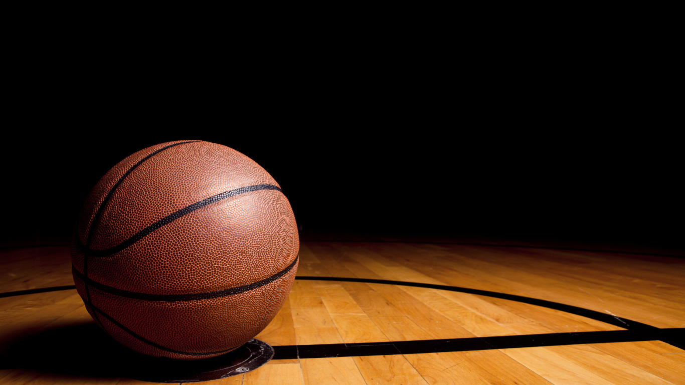 A basketball on a wooden gym floor with black lines, illuminated in a dark setting.