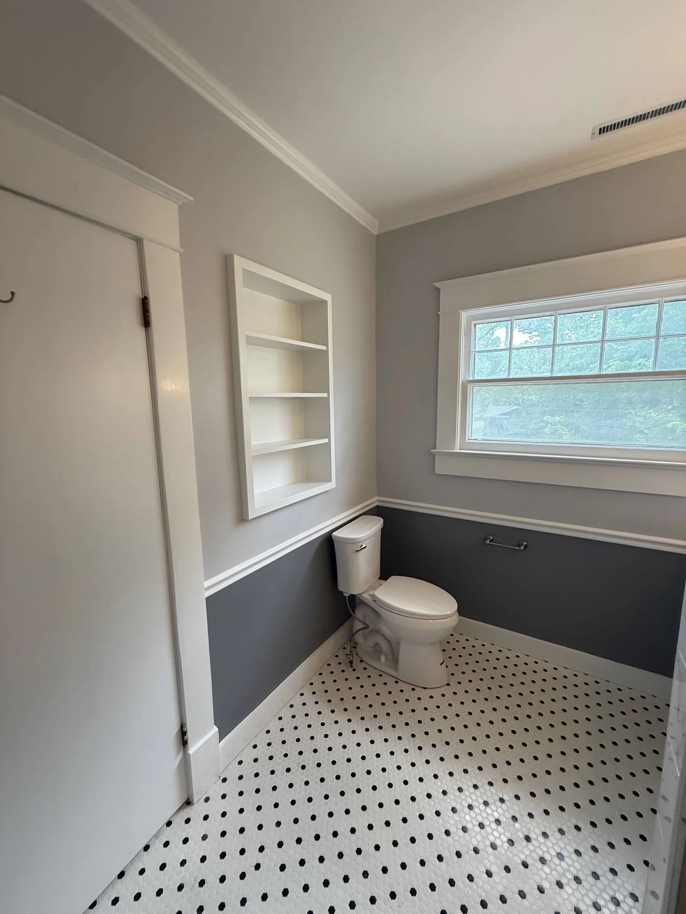 A small bathroom with a white toilet, a window with a view of trees, a white wall-mounted shelf, and a two-tone wall with light gray above and dark gray below, with white trim and black-and-white tiled floor.