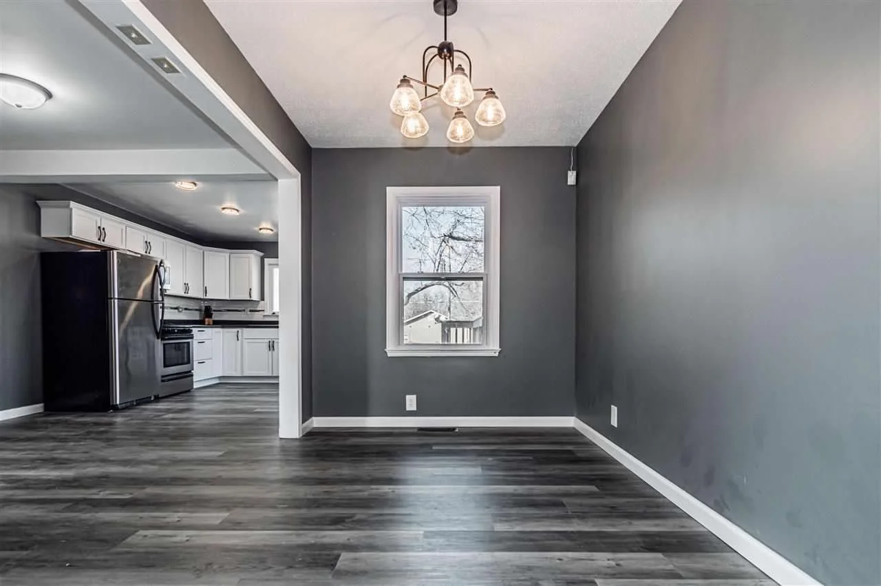 Empty dining area in a house with gray walls, a ceiling light fixture, and a window overlooking a backyard. Adjacent kitchen with white cabinets, stainless steel appliances, and wood flooring.