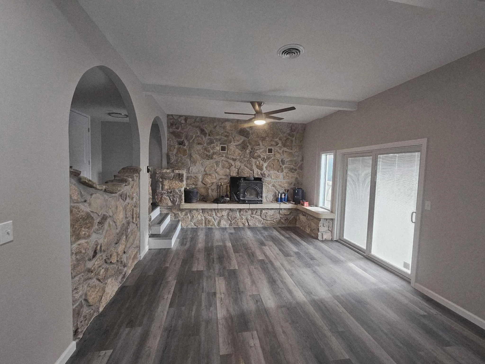 Living room with stone fireplace, gray walls, sliding glass door, wood flooring, ceiling fan, and window.
