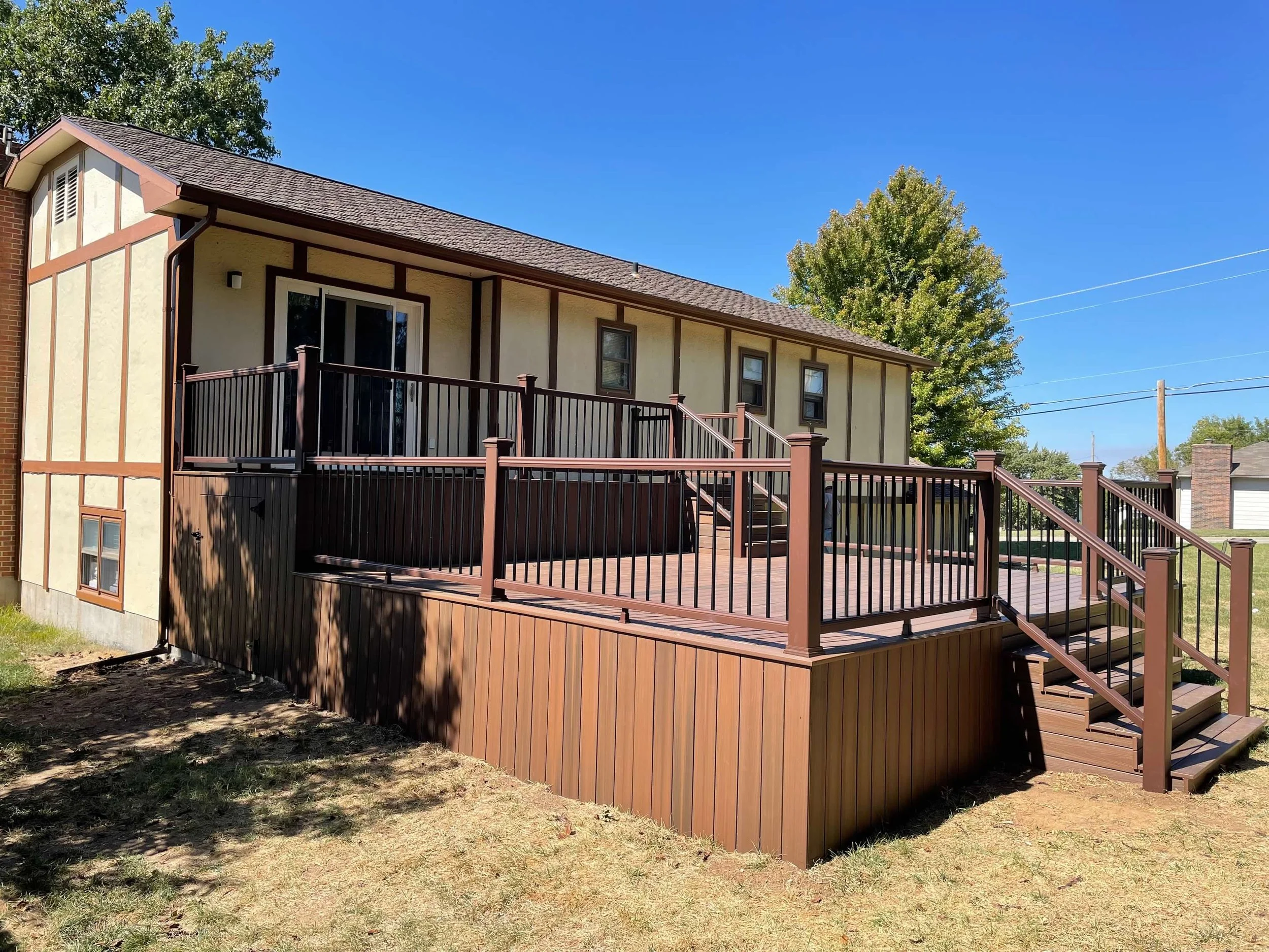 A house with a multi-level wooden deck and railing, stairs leading down to a grassy yard, with trees and a blue sky in the background.
