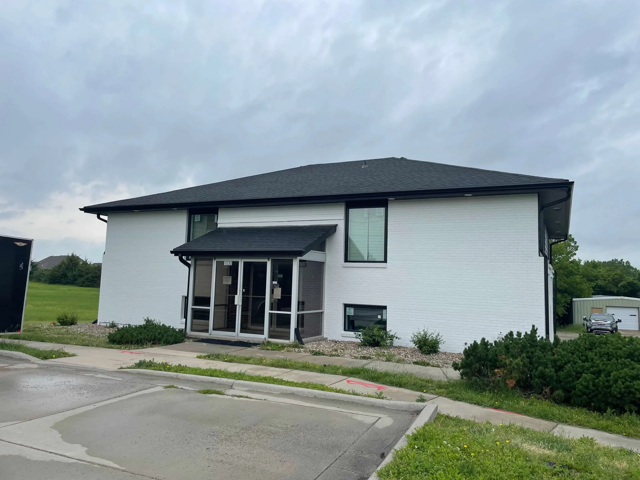 Modern white brick two-story building with black roof and glass entrance doors, surrounded by landscaped yard with small shrubs and bushes, overcast sky.