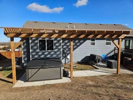 Backyard view of a house with a newly built wooden pergola, a hot tub, and a concrete patio, with a grassy yard and blue sky.