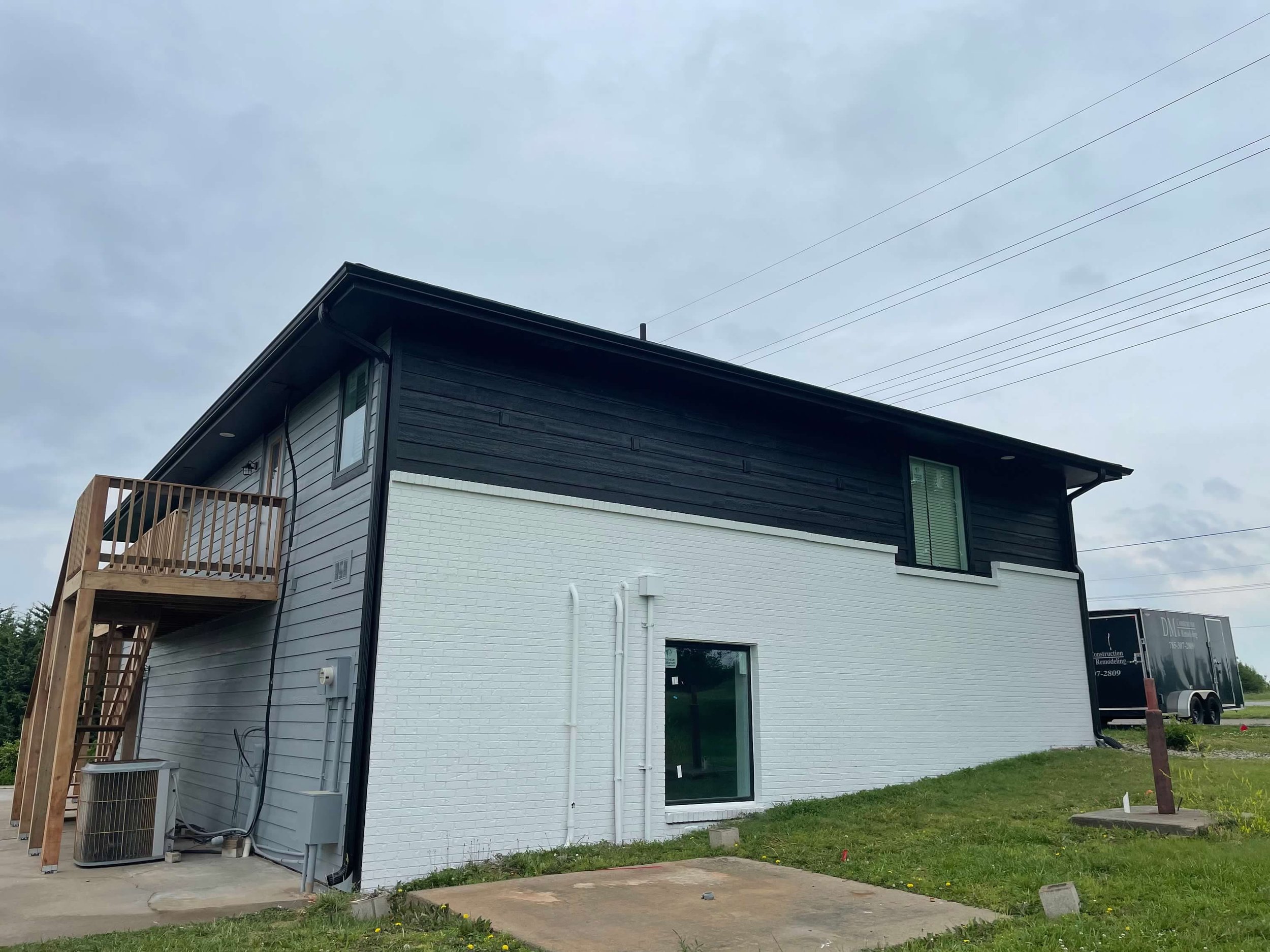 Two-story modern house with black and white exterior, a small wooden balcony on the second floor, and a grassy area in front. A black moving truck is parked nearby.