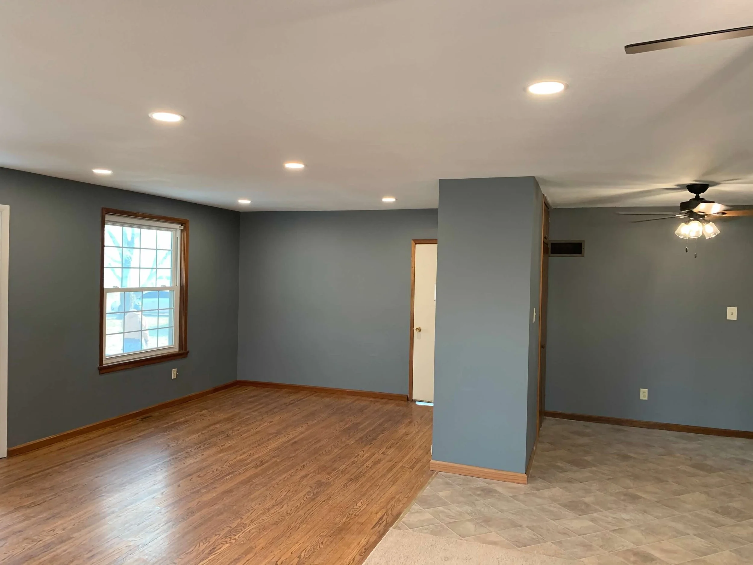 Empty living room with blue walls, hardwood floors, a window, and a ceiling fan with lights.