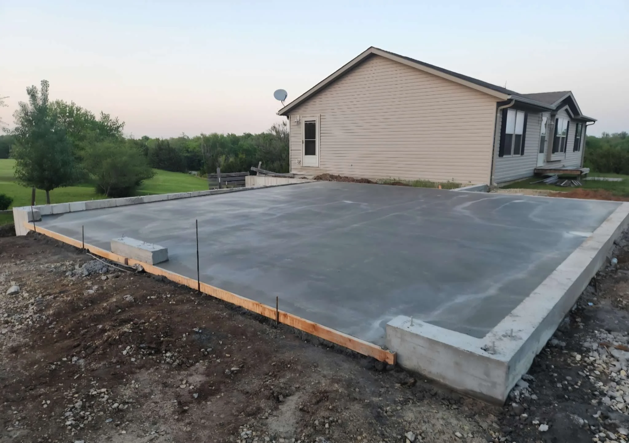 Concrete foundation for a house in construction, with wooden formwork and rebar in place, next to an existing beige house with vinyl siding, windows, and a satellite dish, on a grassy area with trees in the background.