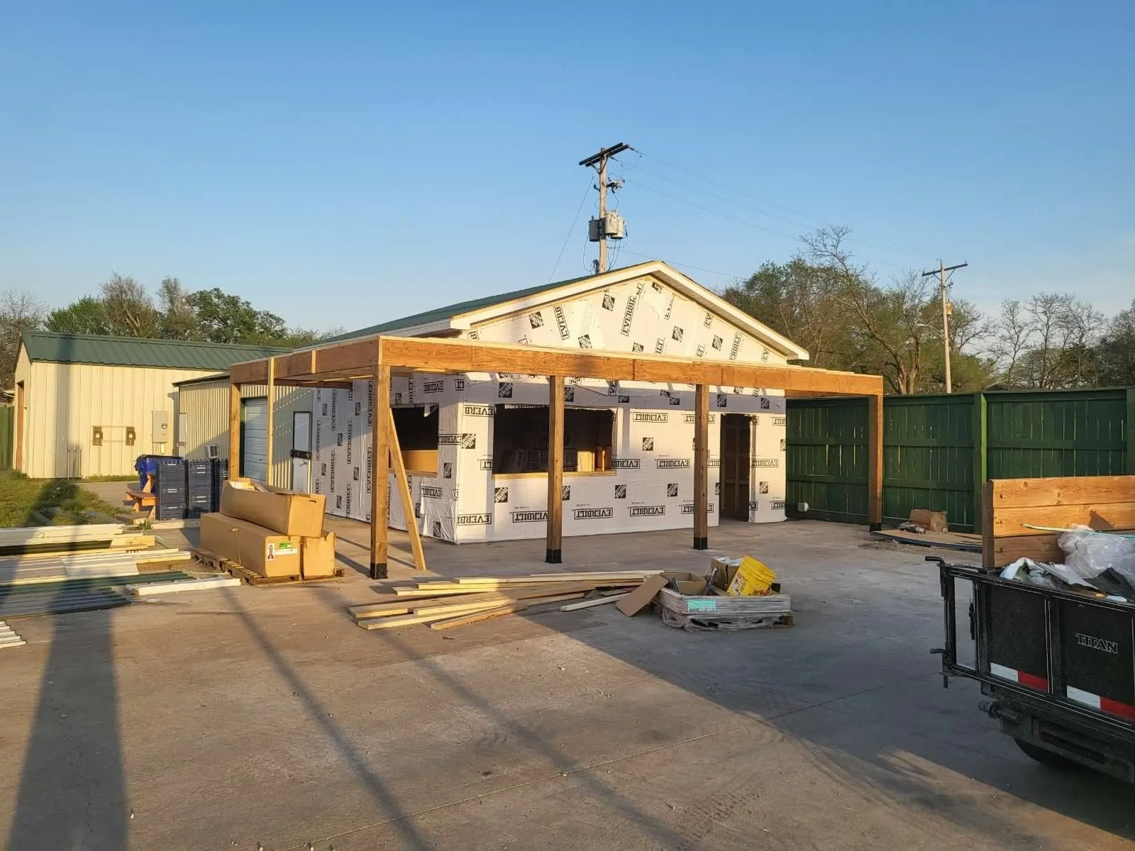Construction site of a small building with a green fence in the background, showing a partially built wooden structure with building materials scattered around, including lumber, boxes, and tools, during daylight.