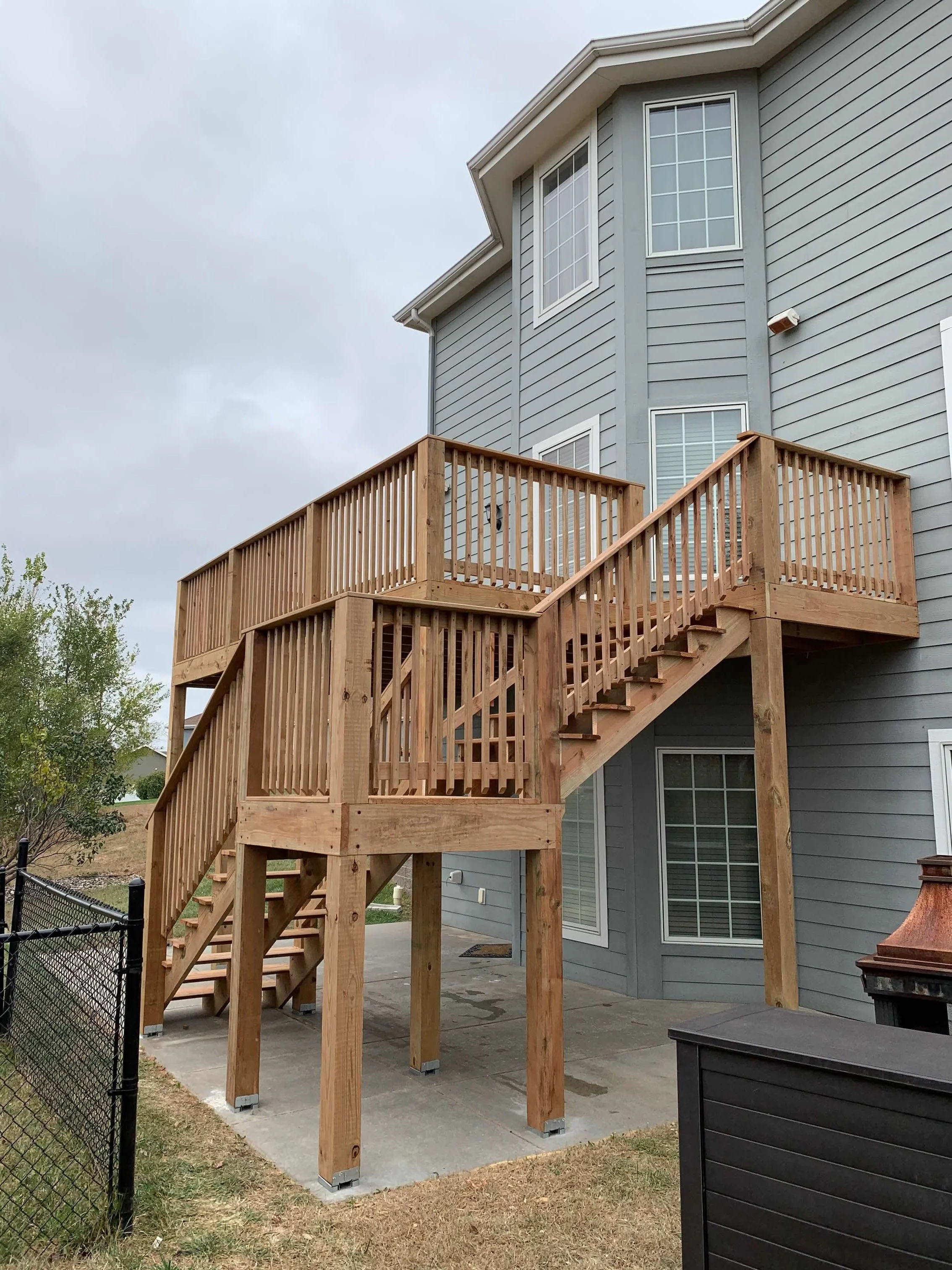 New wooden outdoor staircase and deck attached to the back of a gray house with multiple windows.