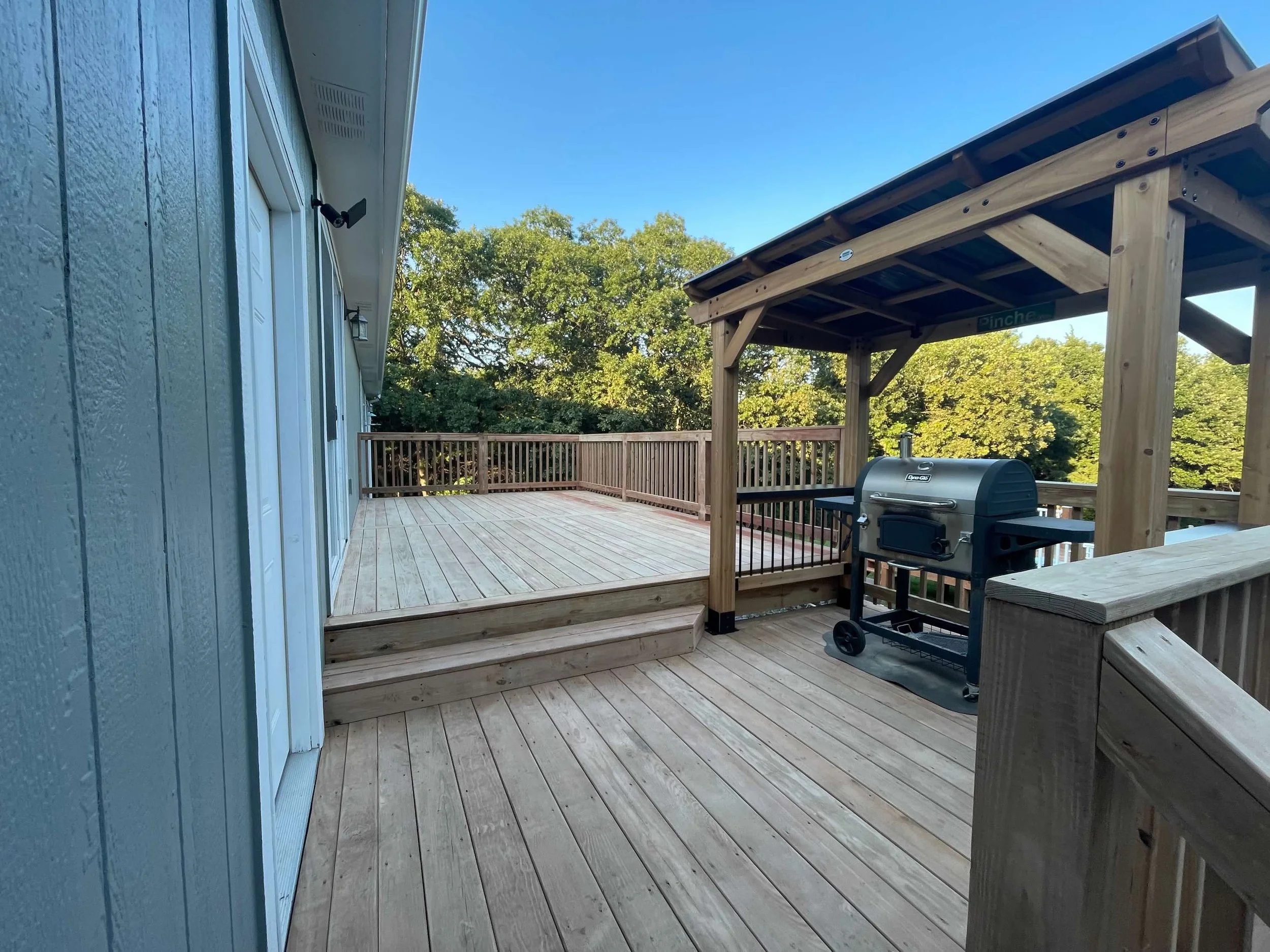 A spacious wooden outdoor deck with a grill under a covered shelter and surrounded by a railing, overlooking green trees during the daytime.